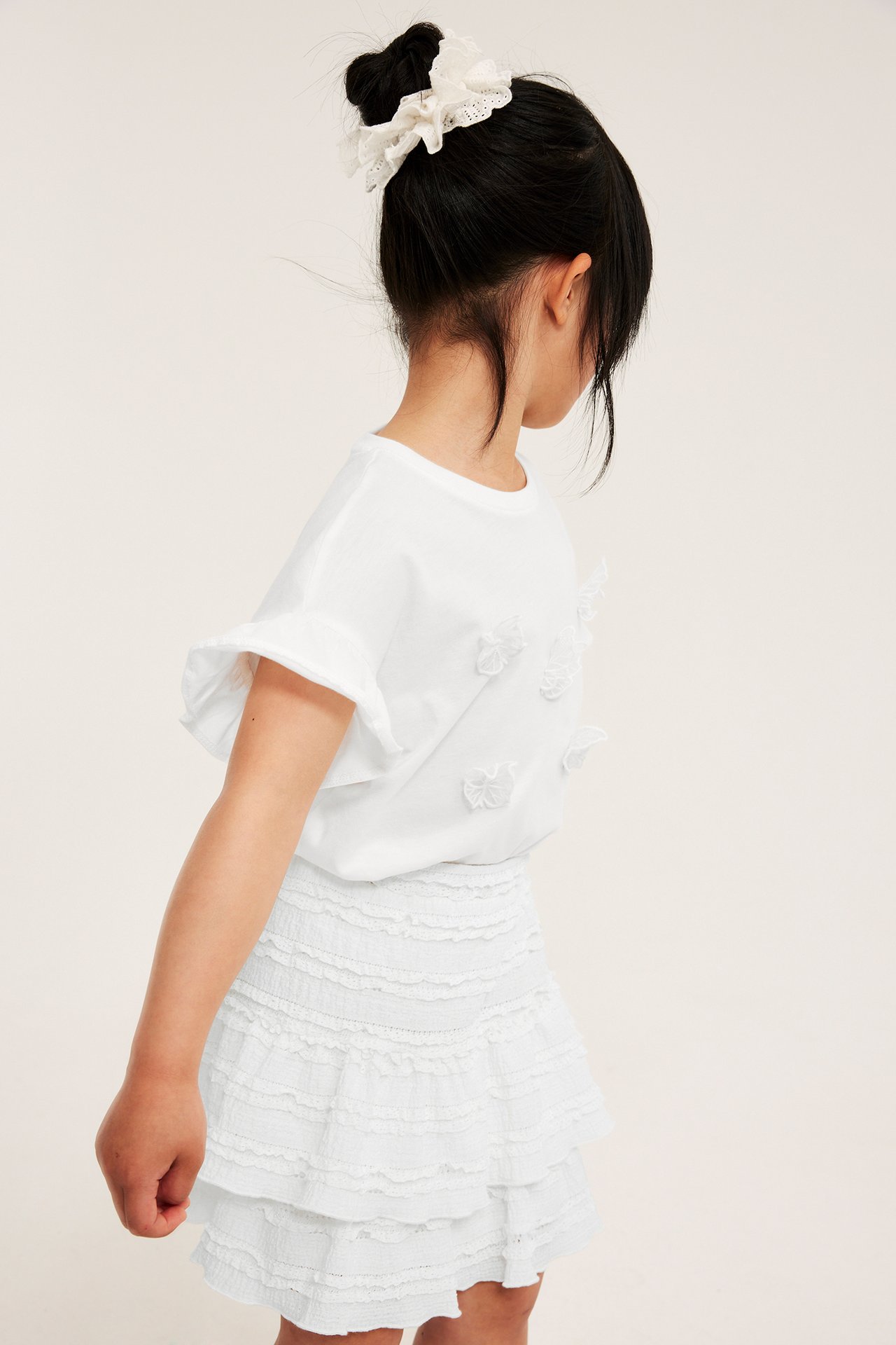 Sidovy av ett barn i en kort offwhite spetsvolangkjol, en vit t-shirt och en spetssnodd.