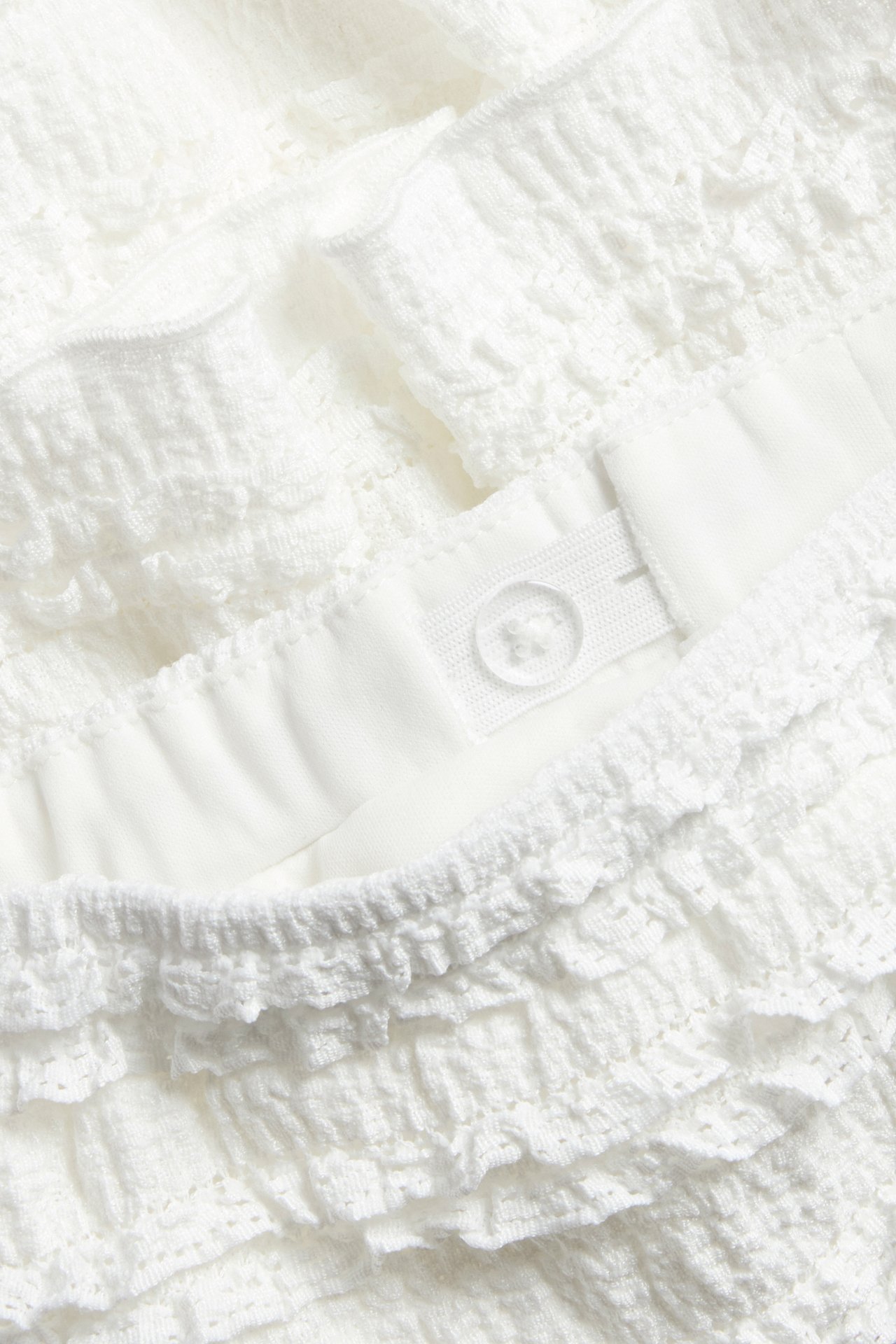 Lähikuva luonnonvalkoisesta lyhyestä pitsirypytyshameesta, jossa on joustava, säädettävä vyötärönappi.