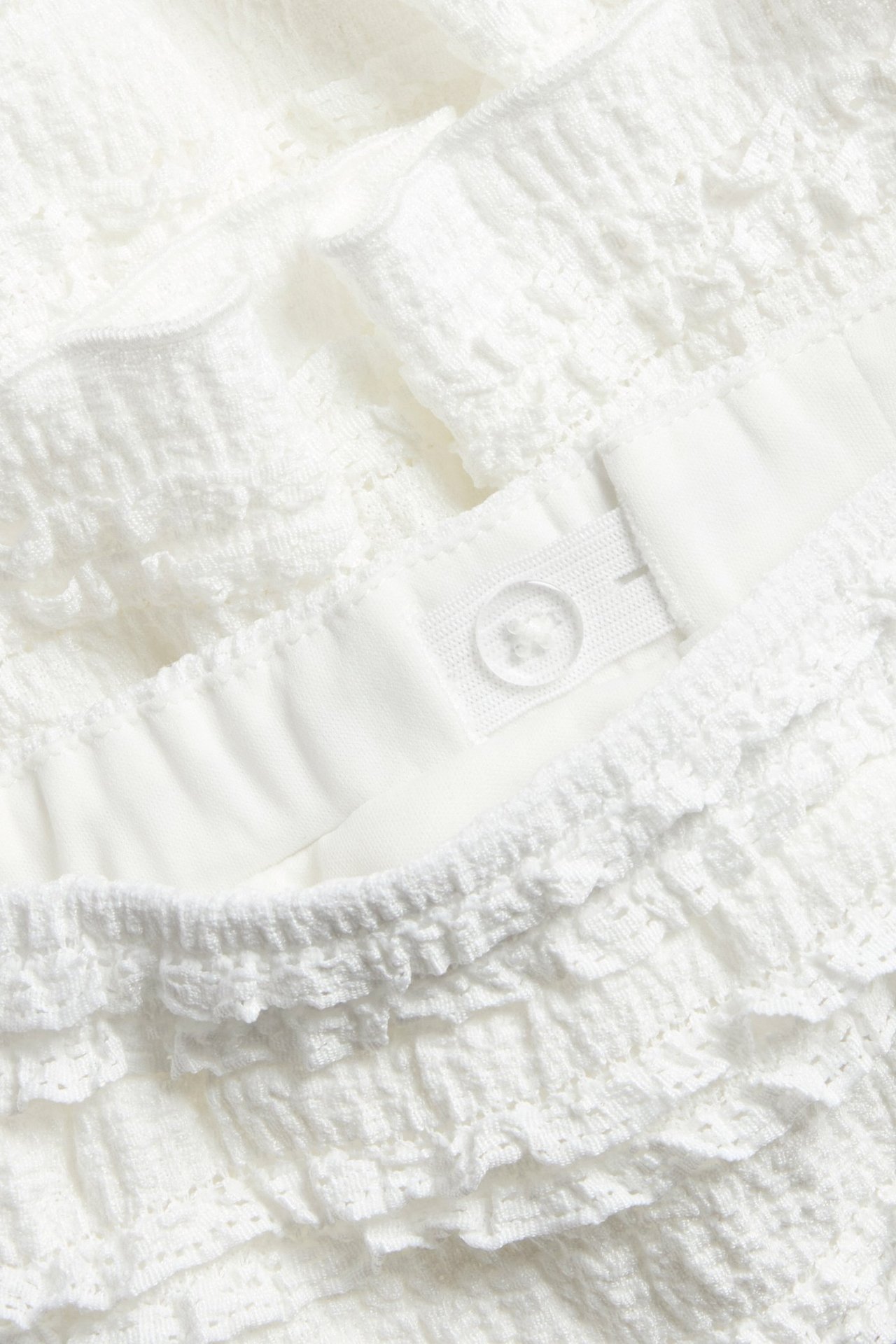 Närbild på en kort, offwhite spetsvolangkjol med resår och justerbar midjeknapp.