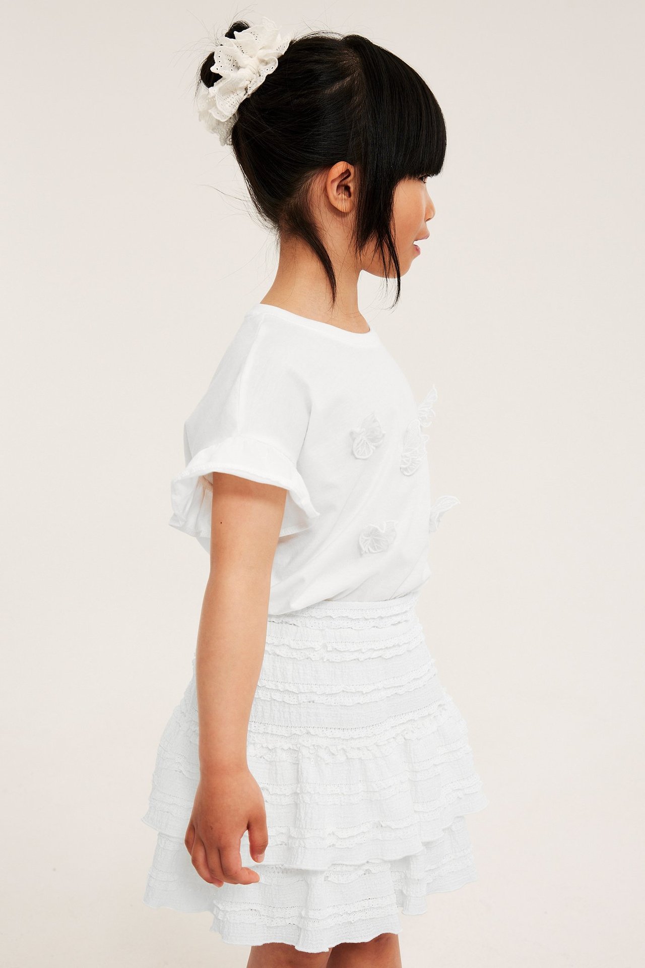 Sidovy: Barn i benvit volangkjol i spets, vit t-shirt med volangärmar och vit spetssnodd.