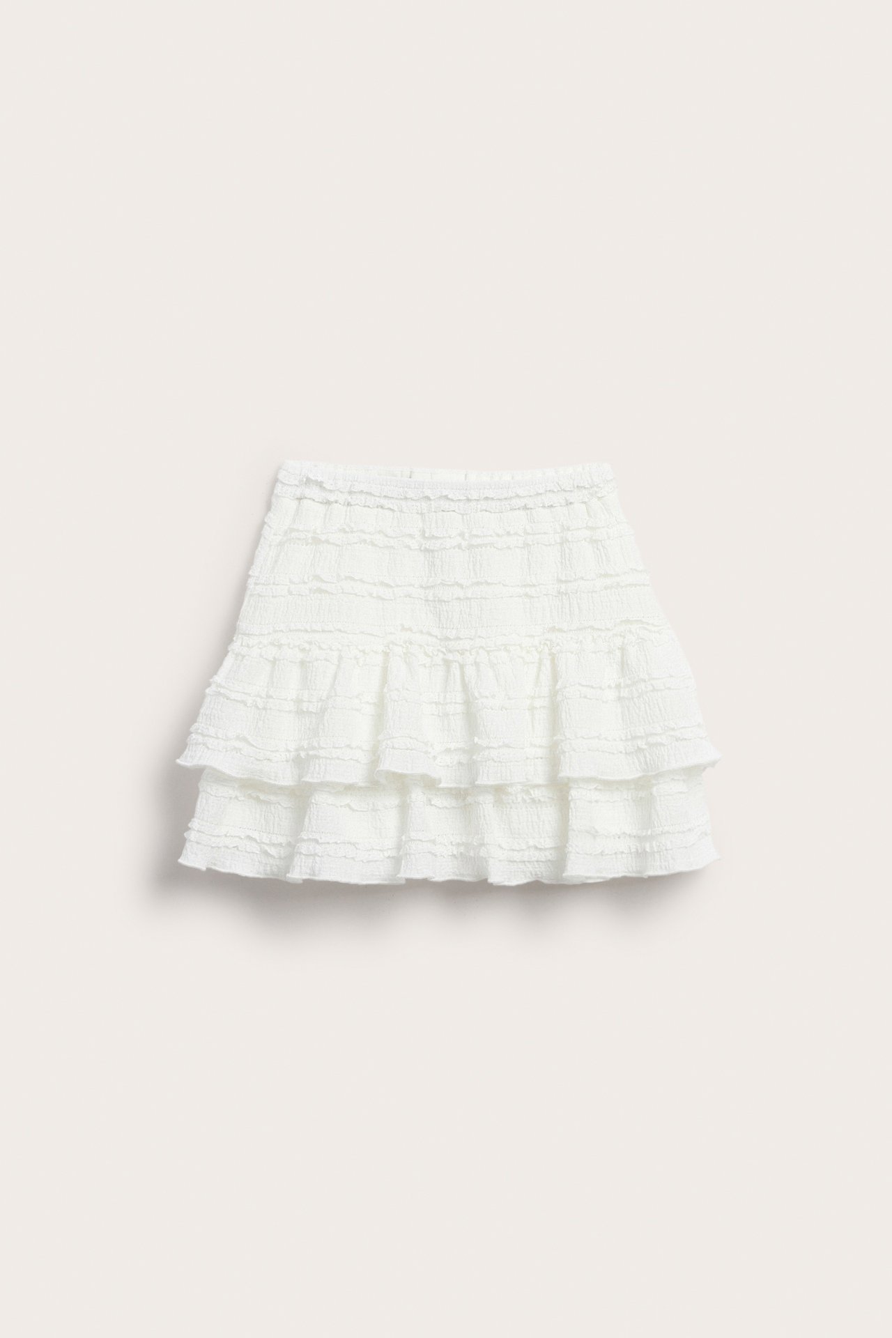 Edestäpäin, lyhyt luonnonvalkoinen röyhelöpitsihame, jossa on joustava, säädettävä vyötärö.