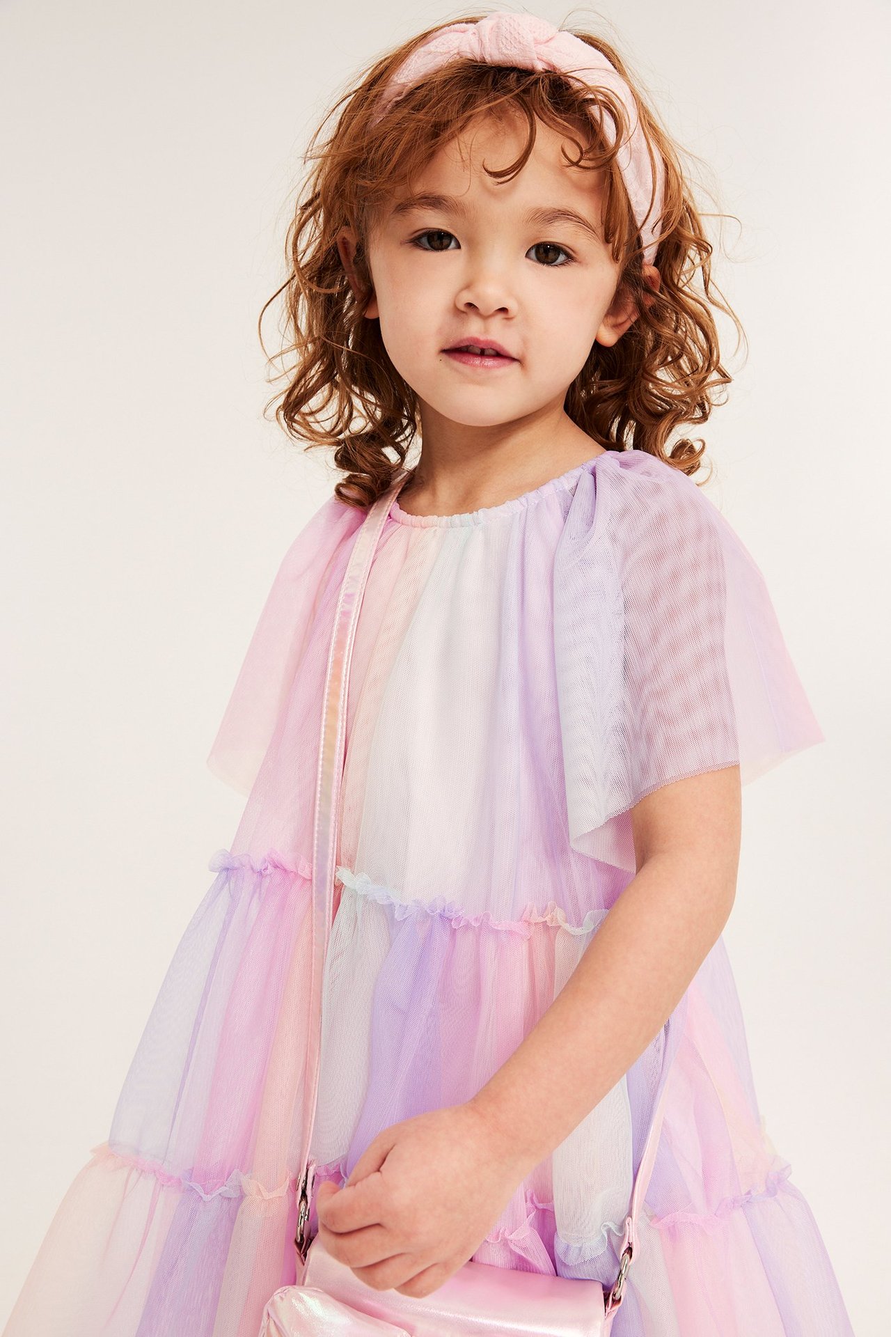 Edestä, lapsi sateenkaarenvärisessä tyllimekossa, vaaleanpunainen rusettipanta ja pitää vaaleanpunaista metallilaukkua.