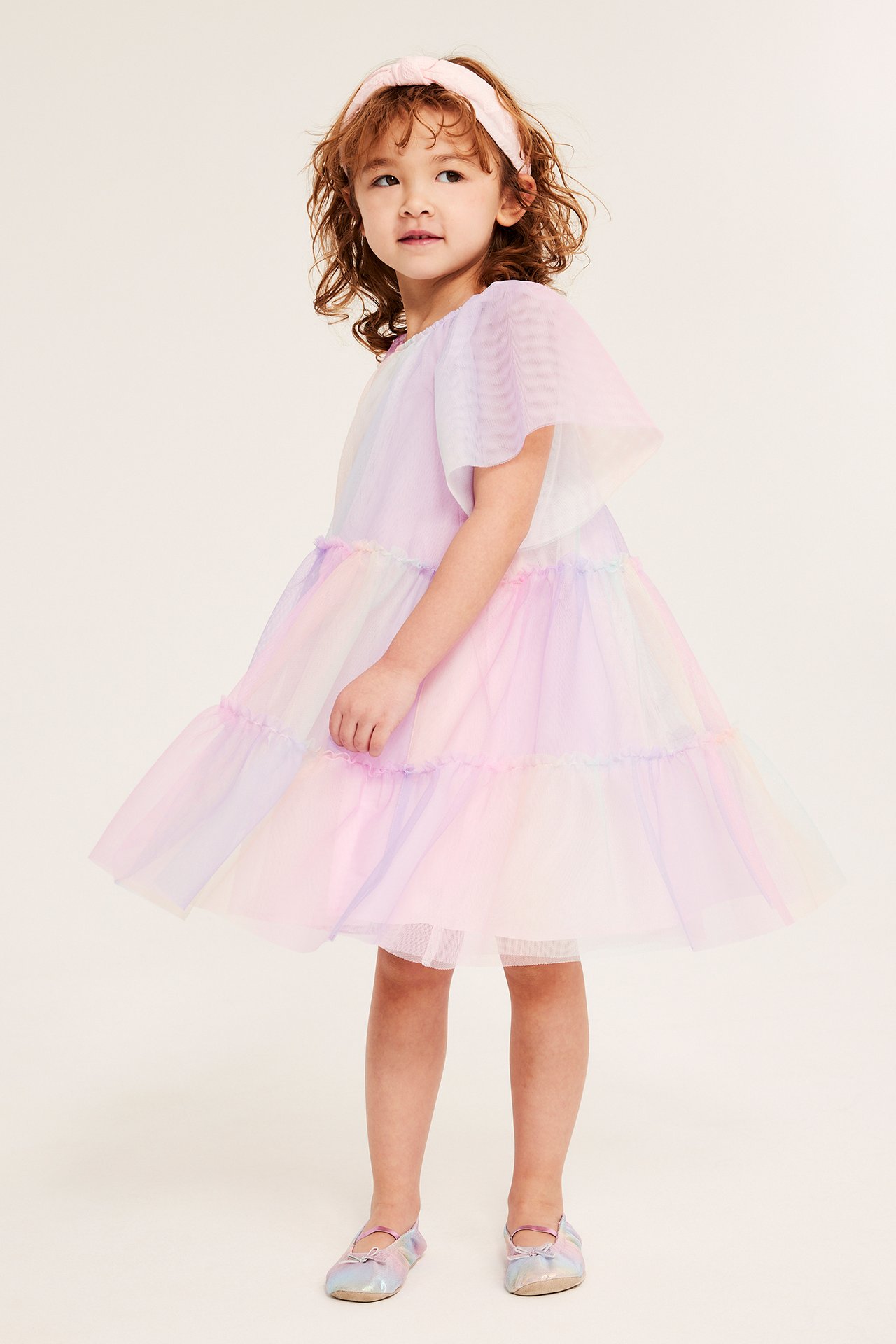 Edestäpäin, lapsi yllään sateenkaarenvärinen runsas tyllimekko, vaaleanpunainen päänauha ja hohtavat ballerinakengät.