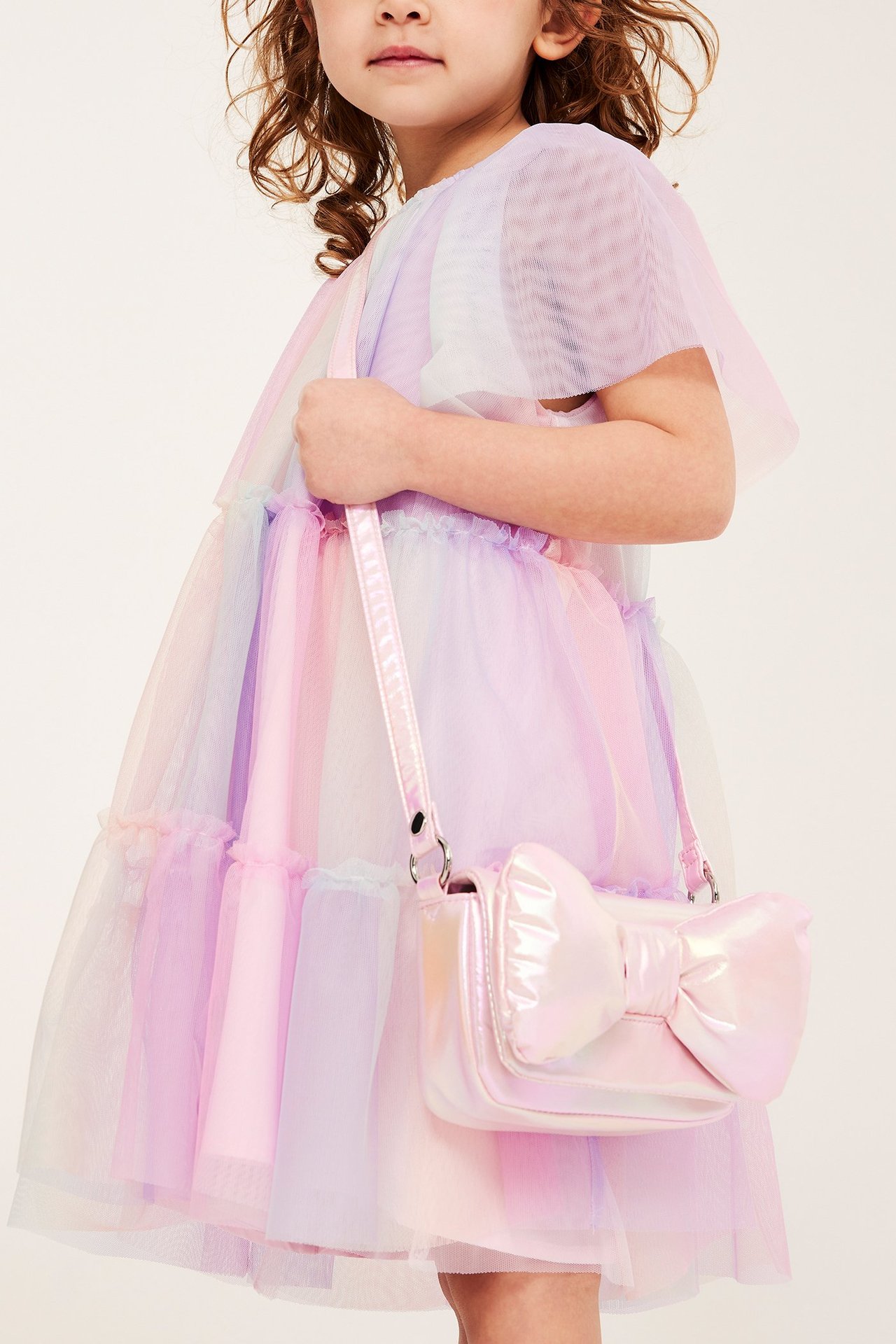 Lapsi edestä sateenkaarenvärisessä kerrostyllimekossa liehuvahihoilla, pitäen vaaleanpunaista helmiäisrusettilaukkua.