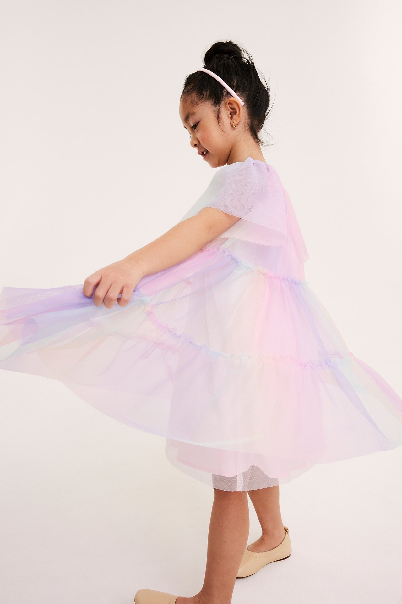 Sidovy av ett barn som snurrar en voluminös regnbågsfärgad tyllklänning, med rosa hårband och beiga platta skor.