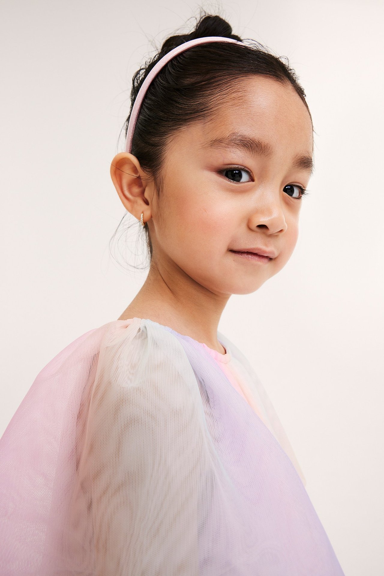 Lähikuva sivulta lapsesta sateenkaarenvärisessä tyllimekossa, vaaleanpunaisessa hiuspannassa ja pienissä rengaskorvakoruissa.