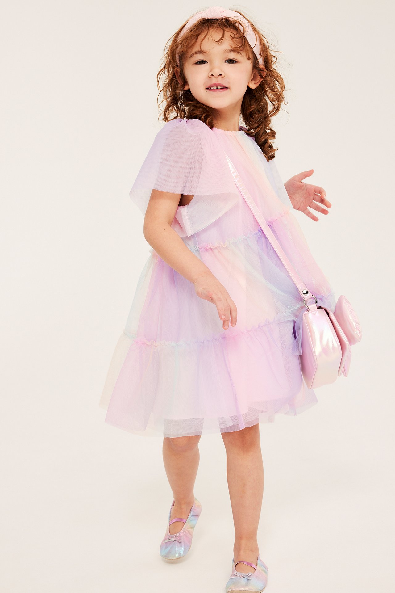 Edestä: Lapsi sateenkaarenvärisessä tyllimekossa, vaaleanpunainen panta, hohtava laukku ja ballerinat.