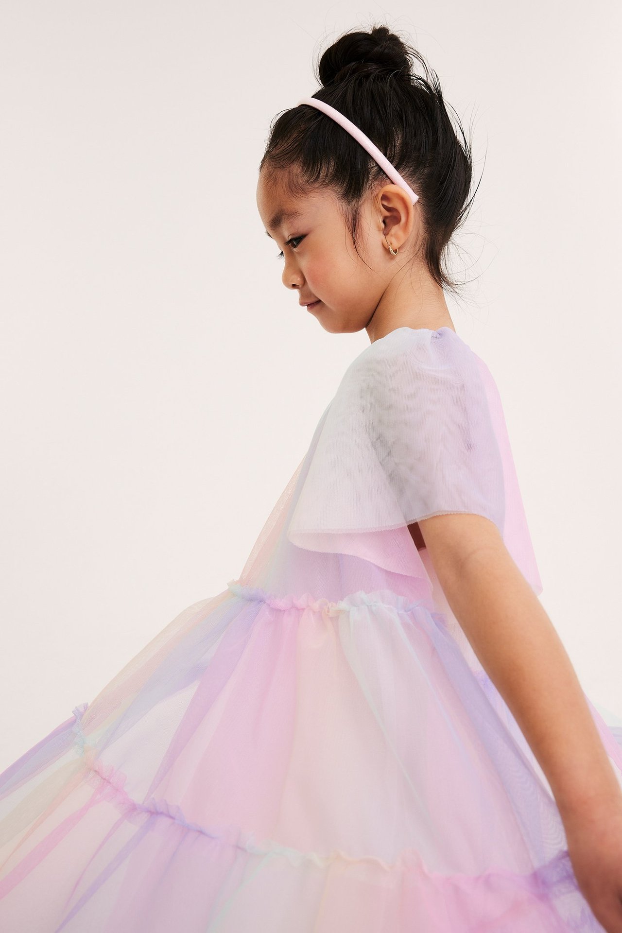 Sivukuva lapsesta väljässä, pastellisateenkaarenvärisessä tyllimekossa ja vaaleanpunaisessa pannassa.