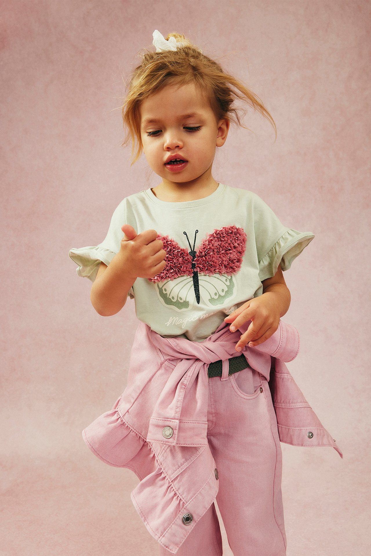 Edestä: Lapsi yllään vihreä puuvillainen röyhelö-T-paita vaaleanpunaisella perhosella, vaaleanpunaiset housut, ja sidottu pinkki takki.