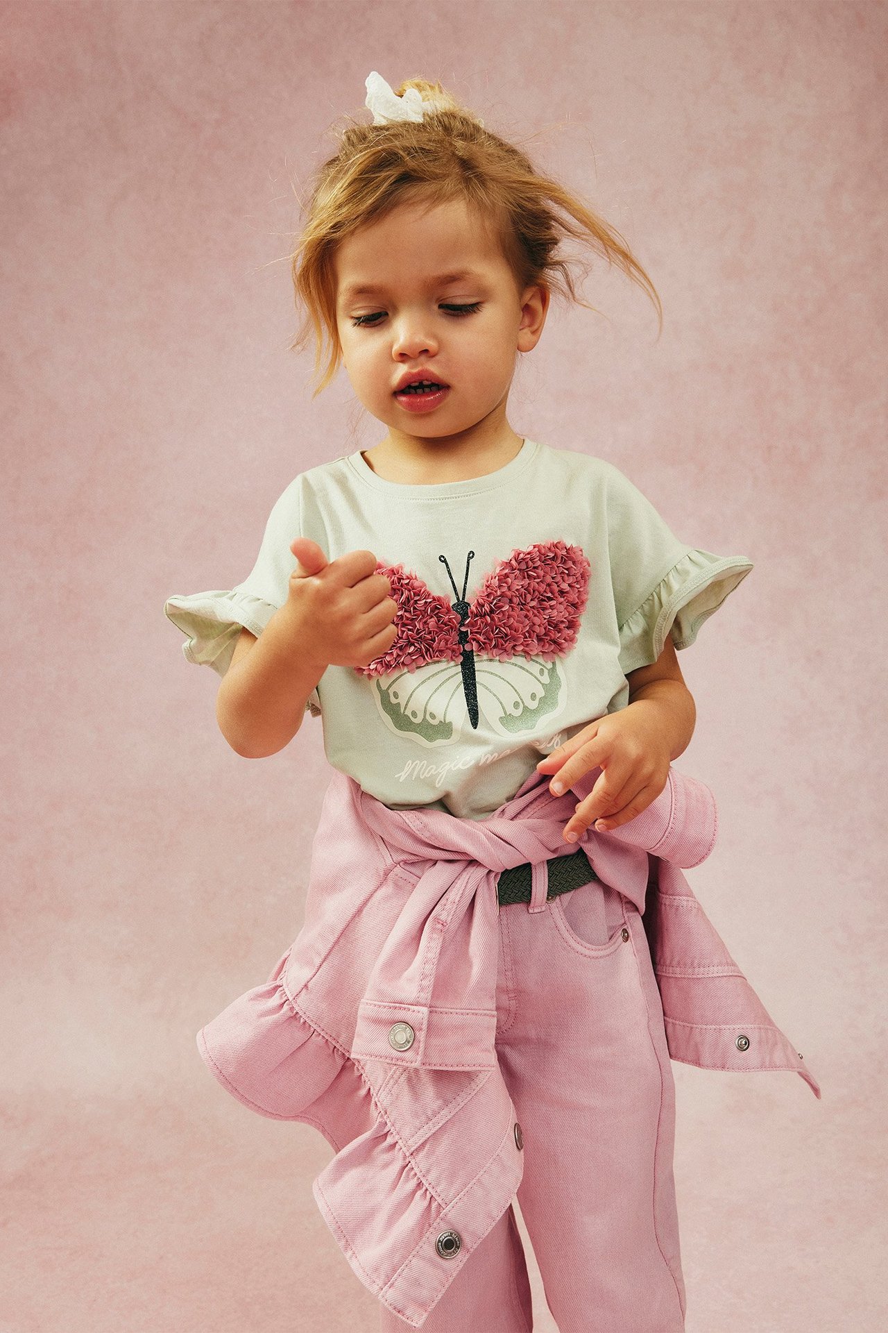 Widok z przodu: Dziecko w zielonej bawełnianej koszulce z falbankami i różowym motylem, różowe spodnie i zawiązana różowa kurtka.