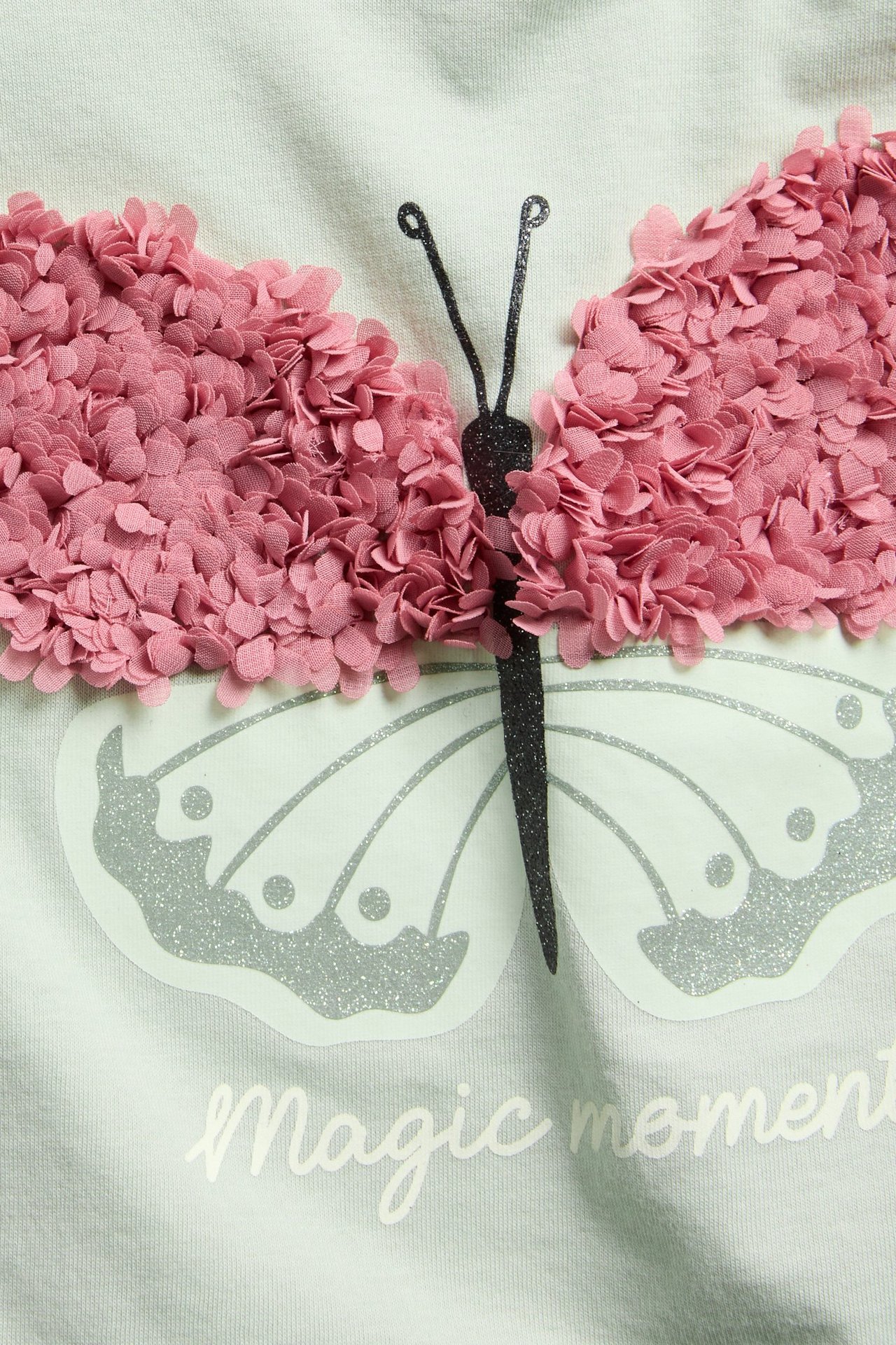 Närbild framifrån på en ljusgrön barnt-shirt med stort rosa 3D-volangfjärils- och glittertryck.