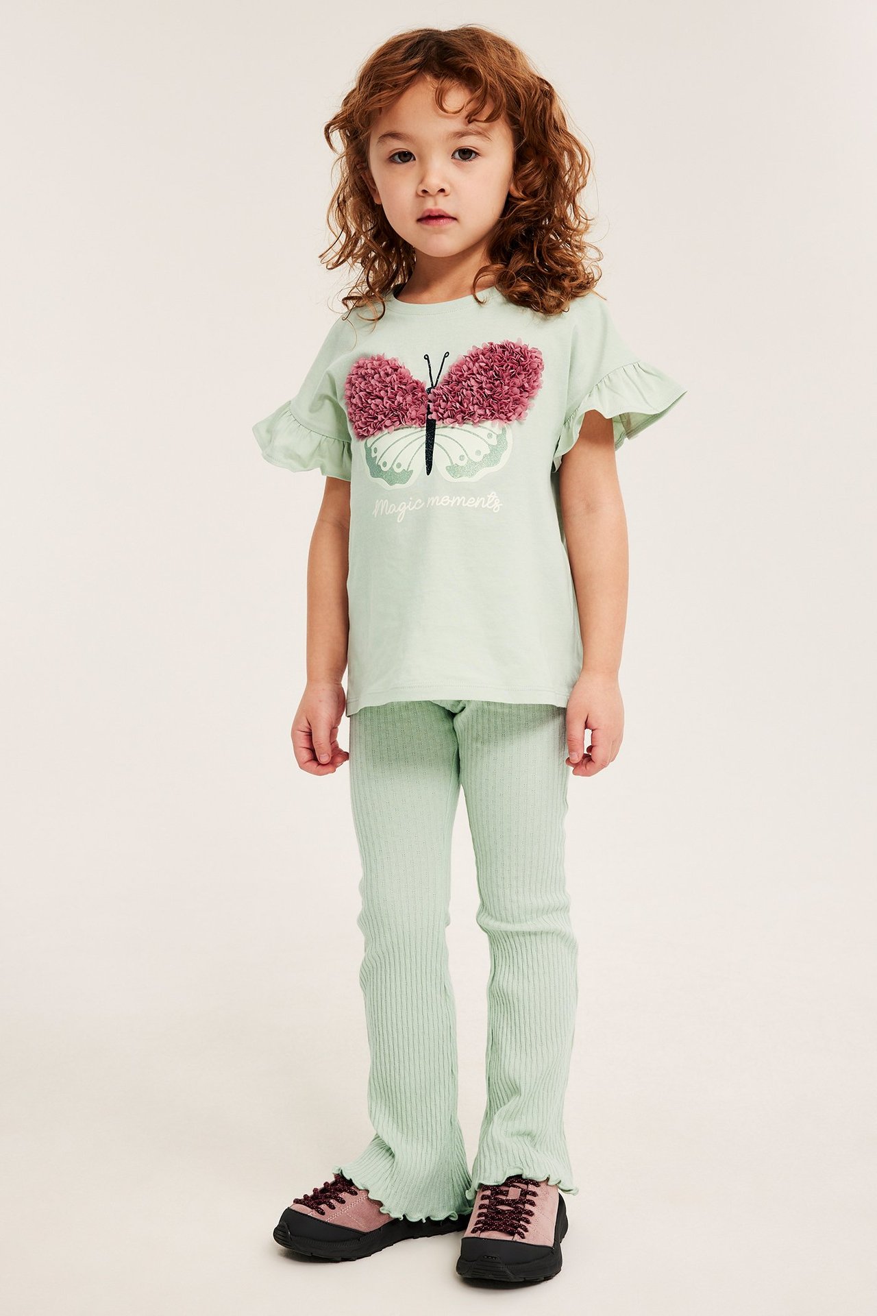 Widok z przodu: Dziecko nosi zielone prążkowane legginsy dzwony, jasnozieloną bluzkę z motylem i różowe buty.
