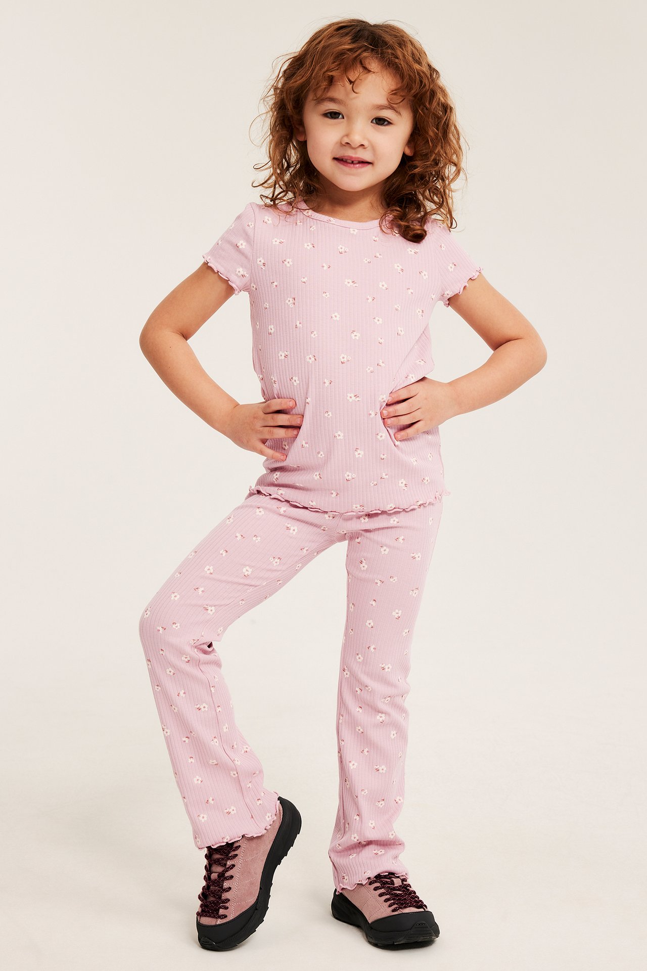 Widok z przodu: Dziecko w jasnoróżowych, kwiecistych, prążkowanych rozszerzanych legginsach, pasującym topie i trampkach.