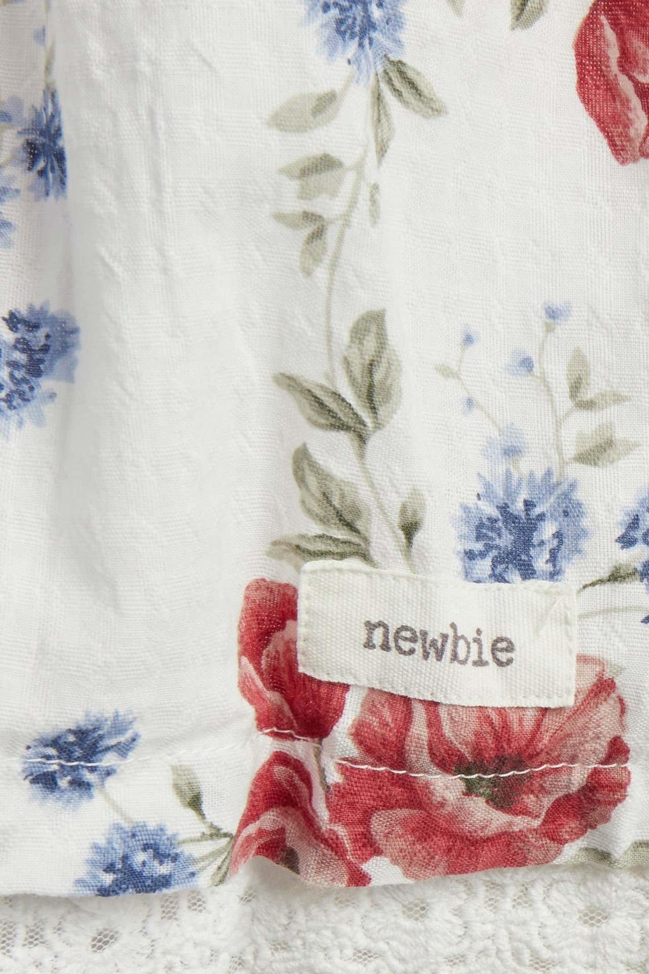 Zbliżenie sukienki w kwiaty, off-white, z czerwonymi i niebieskimi kwiatami, koronką i metką Newbie.
