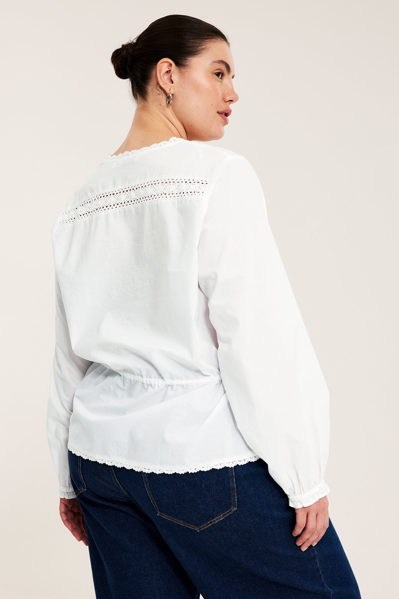 Bakifrån: Kvinna i vit bomullsblus med spets, dragsko i midjan och mörkblå jeans.