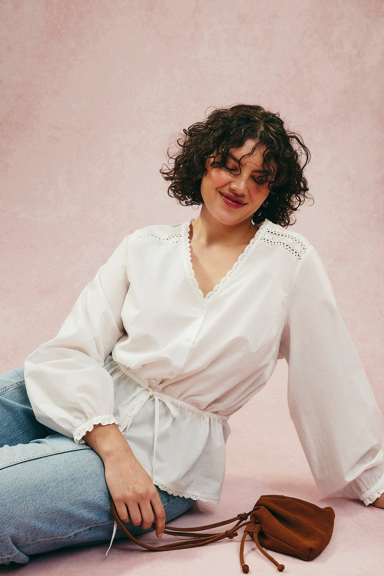 Kvinna bär vit bomullsblus med spets, dragsko i midjan, resår i ärmslut, blå jeans, brun väska.