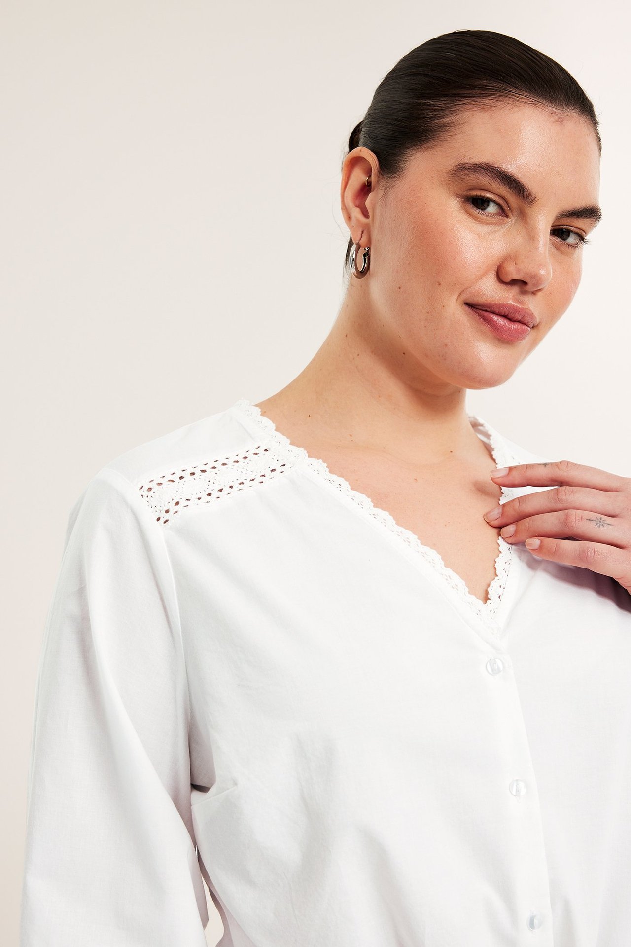 Z przodu zbliżenie: kobieta w białej bawełnianej bluzce z koronkowym dekoltem V, guzikami i kolczykami.