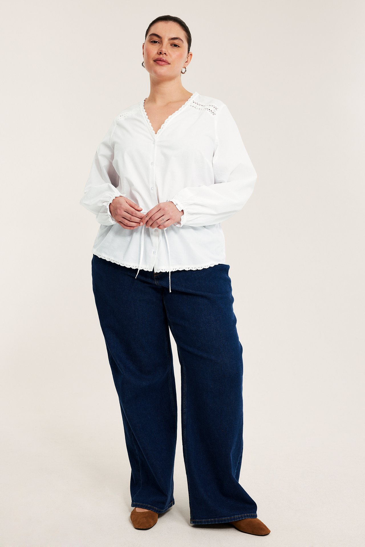 Frontutsikt: Kvinne i hvit bomullsbluse med blonder, snøring, mørkeblå vide jeans og brune flats.