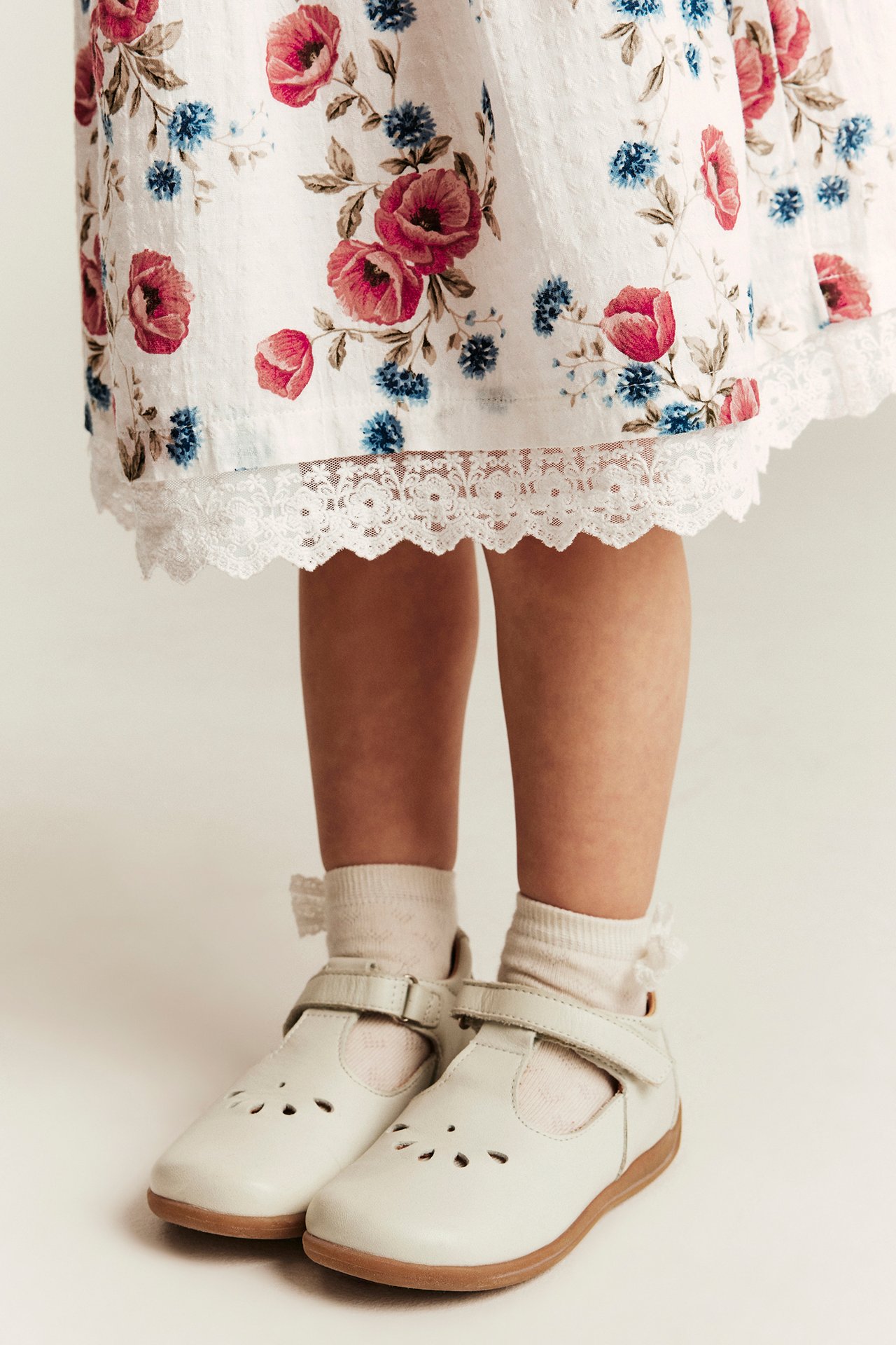 Hvit blomstret kjole med blondekant på barn, hvite sokker og hvite Mary Jane-sko.