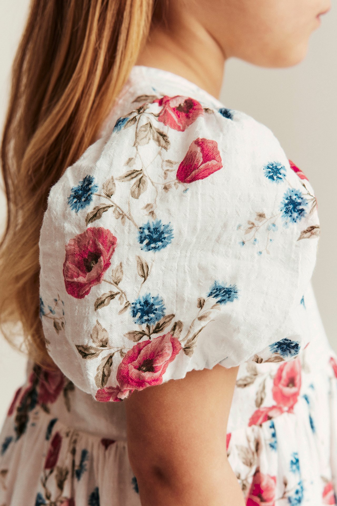 Sidevisning av et barn i en kremfarget blomsterkjole med rosa og blå blomster og ballongermer.