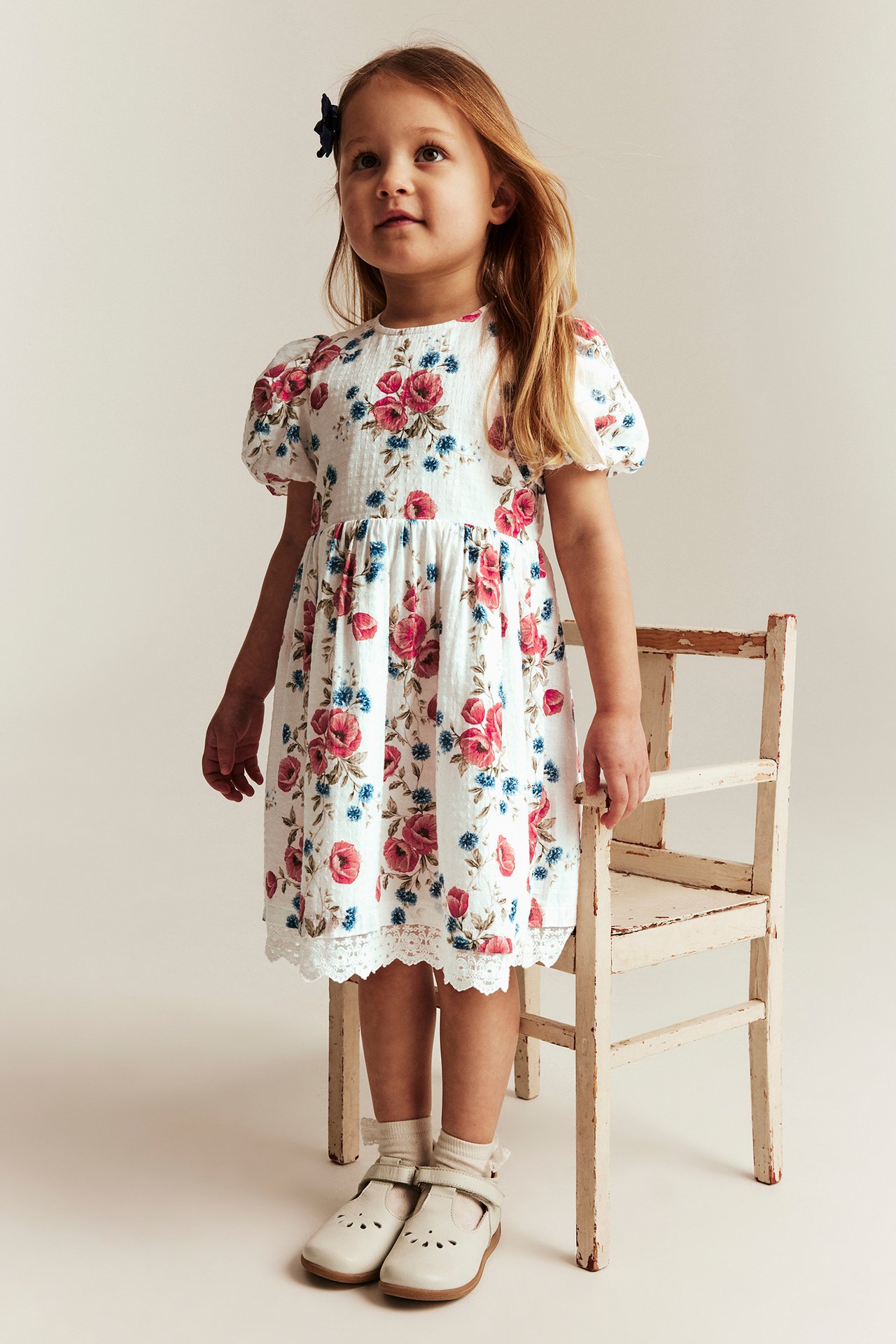 Edestä, lapsi luonnonvalkoisessa kukkamekossa pussihihoilla, pitsireunuksella ja luonnonvalkoisilla kengillä.