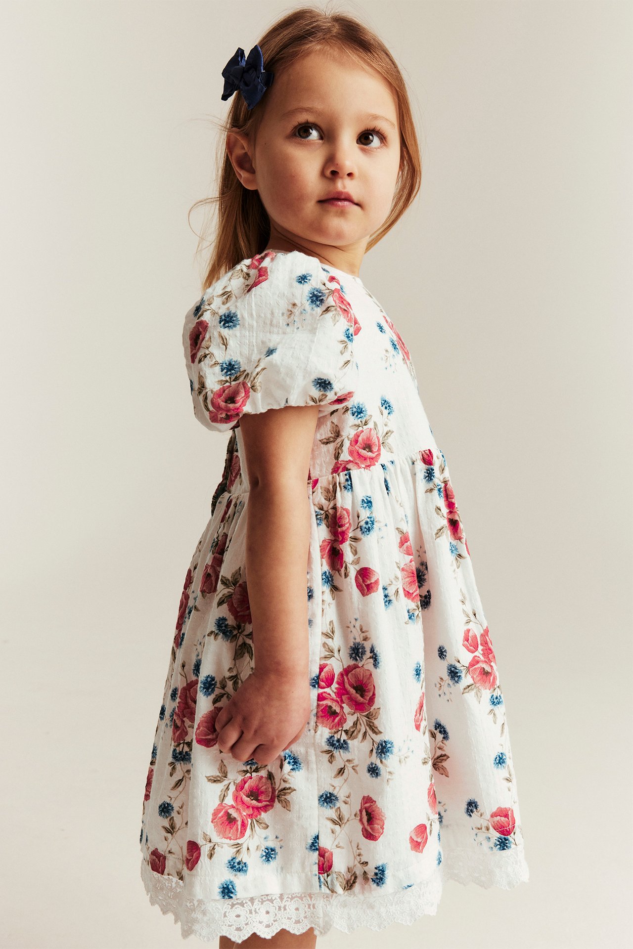 Sivukuva lapsesta yllään luonnonvalkoinen kukkamekko pussihihoilla ja tummansininen hiusklipsi.