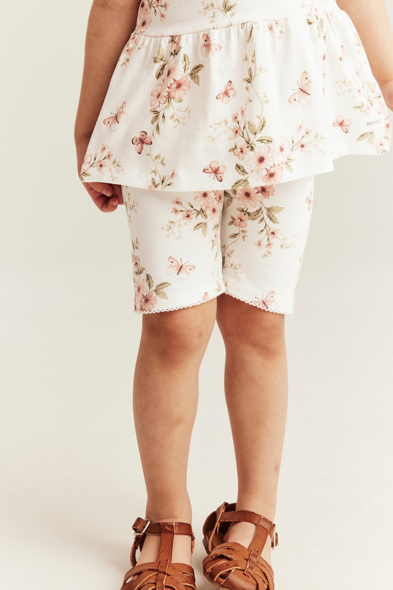 Frontvy: Kid bär offvita blommiga cykelbyxor med picotkant, en matchande topp och bruna sandaler.