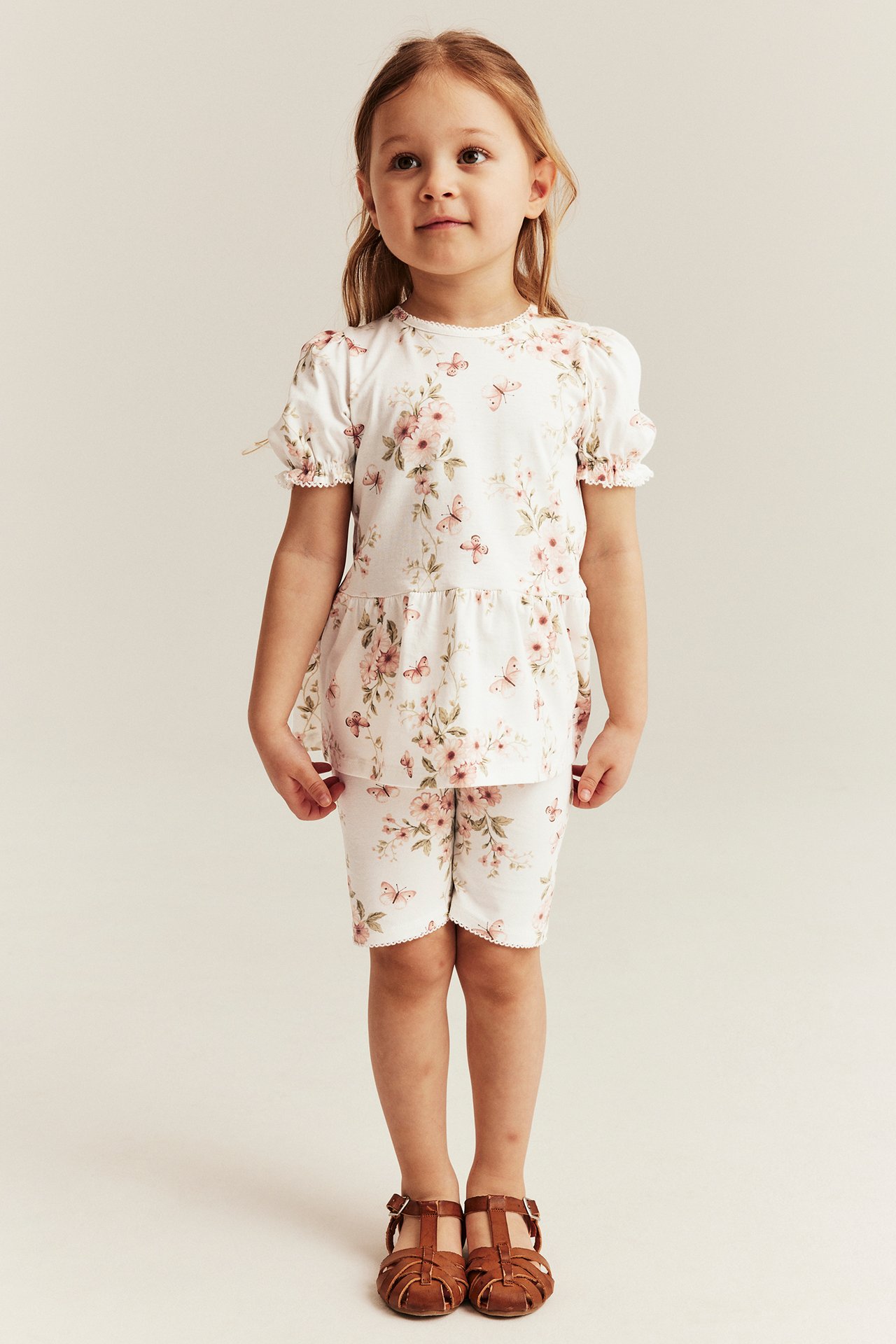 Widok z przodu dziecka w szortach rowerowych ecru z kwiatami i motylami, pasującym topie i brązowych sandałach.