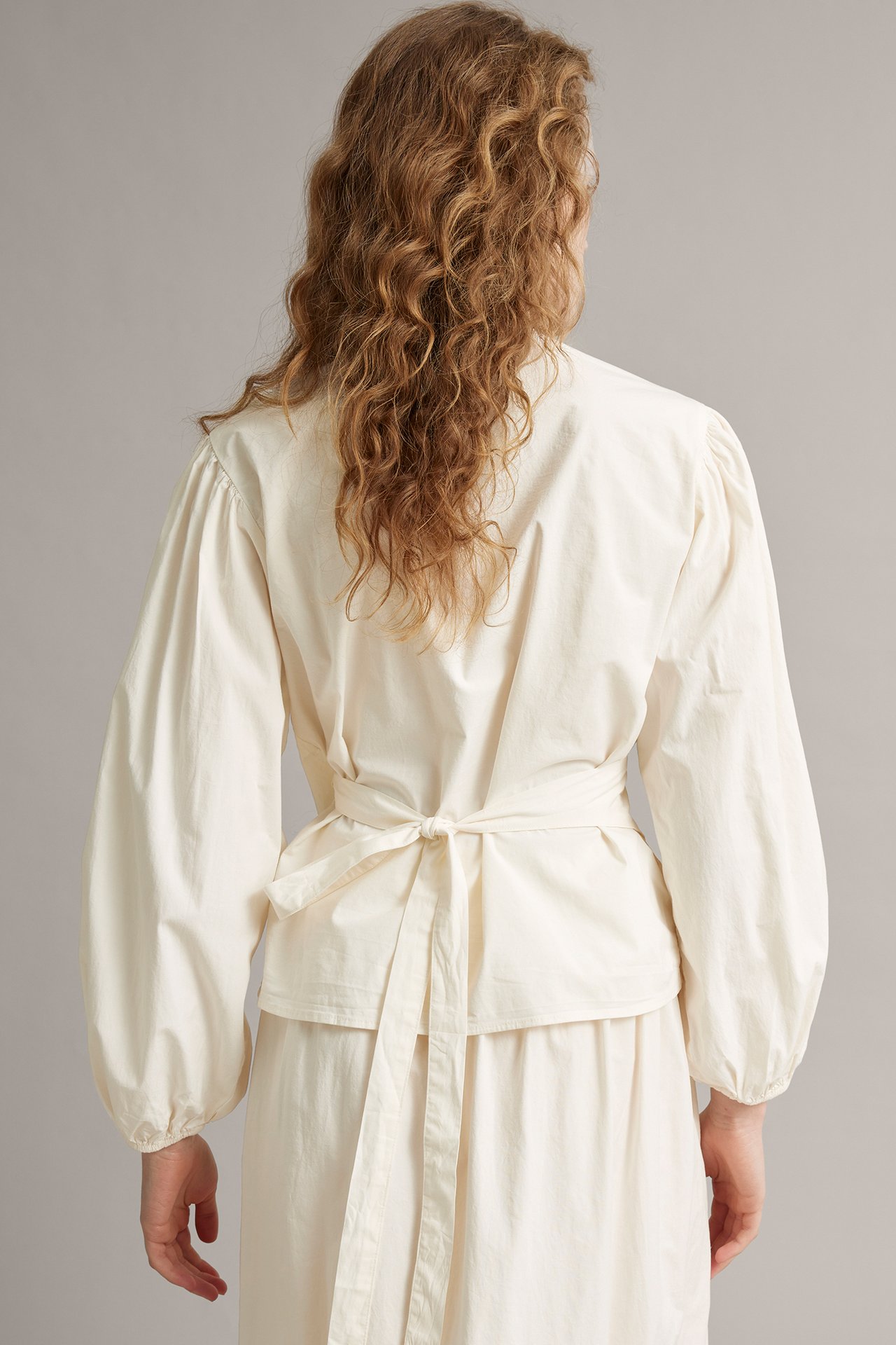 Nainen valkoisessa popliinikietaisupuserossa pussihihoilla, takaa solmittu, ja yhteensopiva hame.