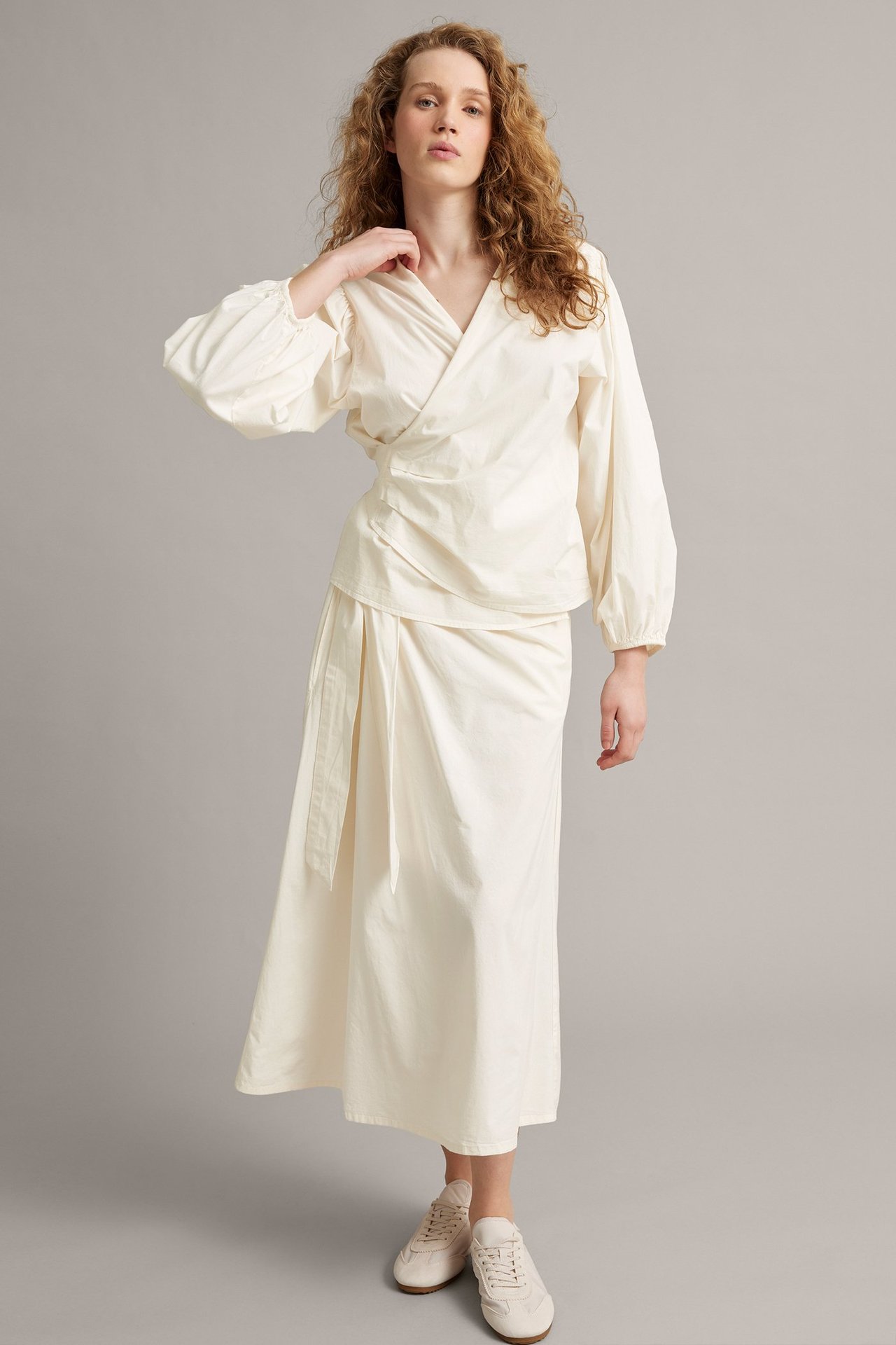 Frontvy av kvinna i cremefärgad omlottblus med vida ballongärmar, matchande midikjol och ljusa sneakers.