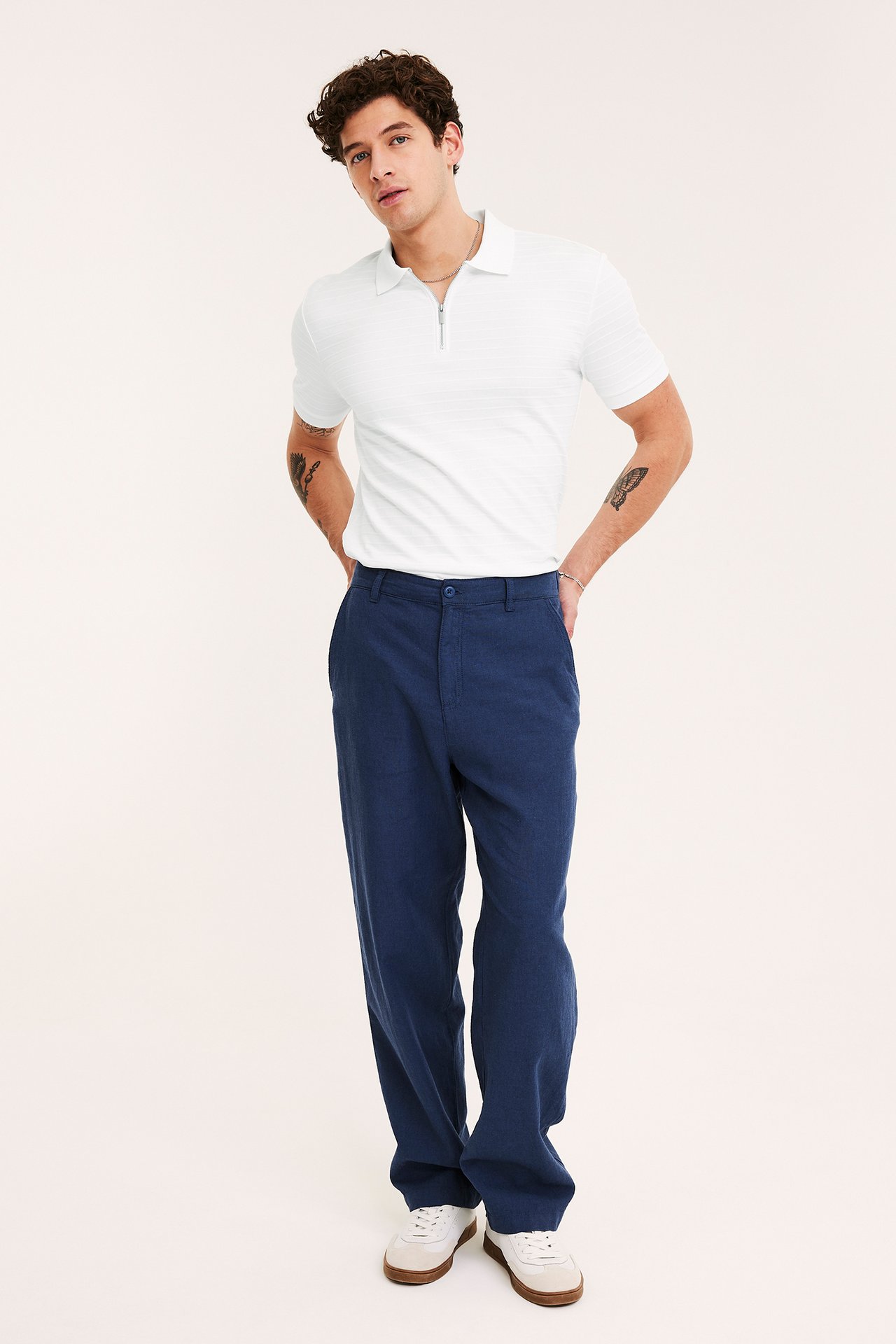 Edestä päin mies valkoisessa lyhythihaisessa vetoketjullisessa pikeepaidassa, tummansinisissä housuissa ja valkoisissa lenkkareissa.