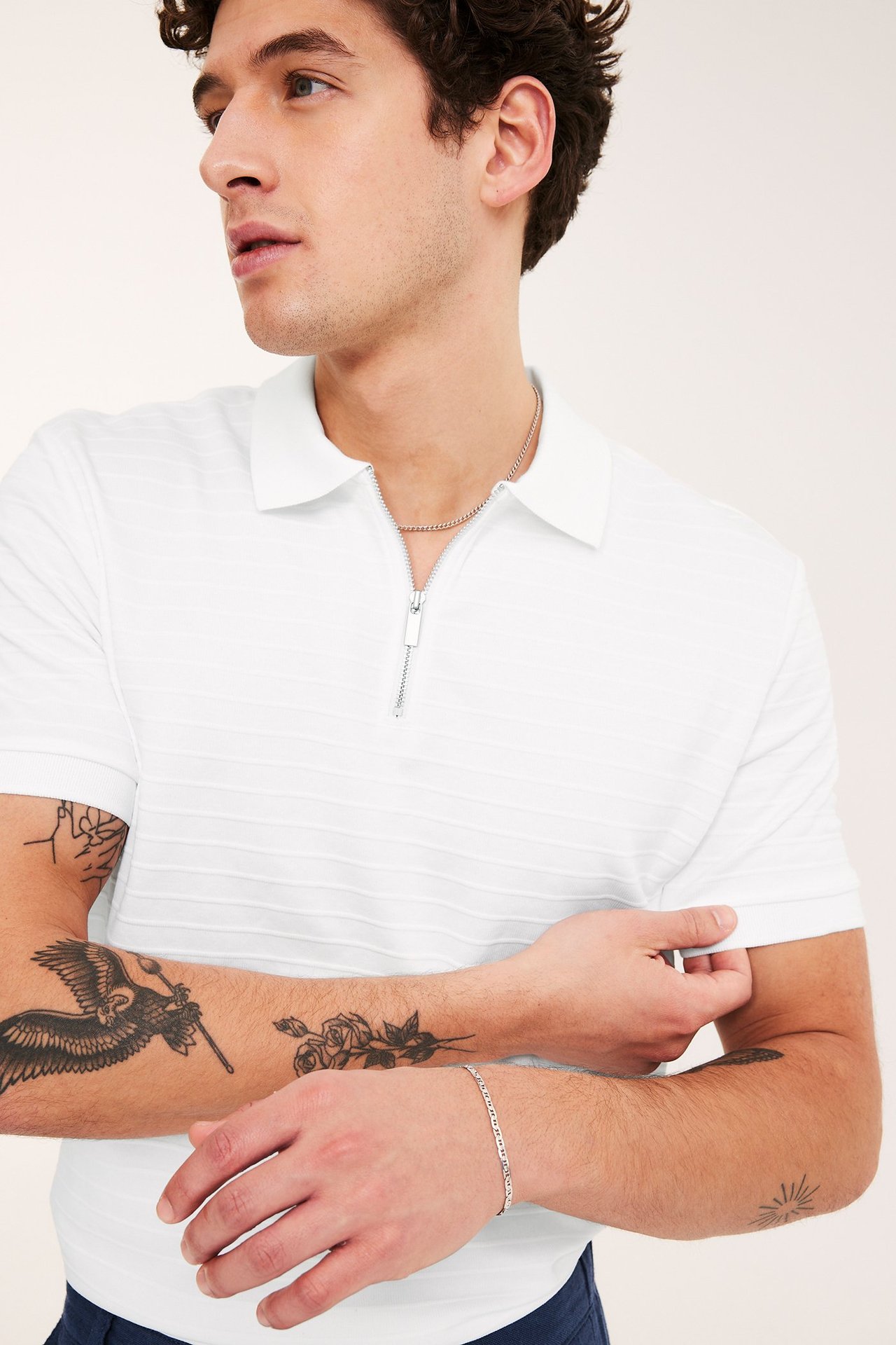 Lähikuva miehestä yllään valkoinen kuvioitu lyhythihainen pikeepaita vetoketjulla, hopeakaulakoru ja rannekoru.