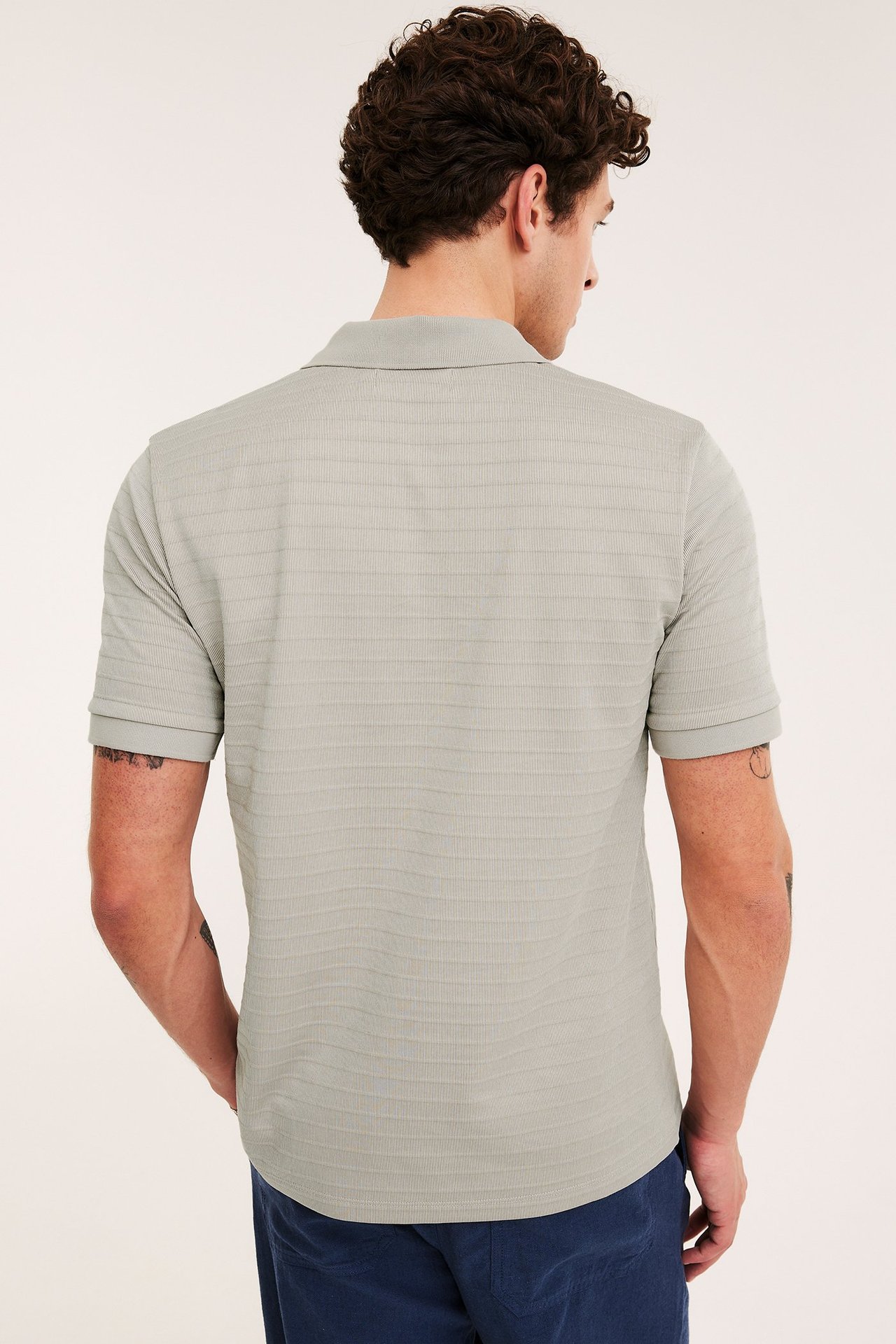 Miehen takakuva beigessä lyhythihaisessa pikeepaidassa, jossa hienostunut ribattu tekstuuri, ja tummansinisissä housuissa.