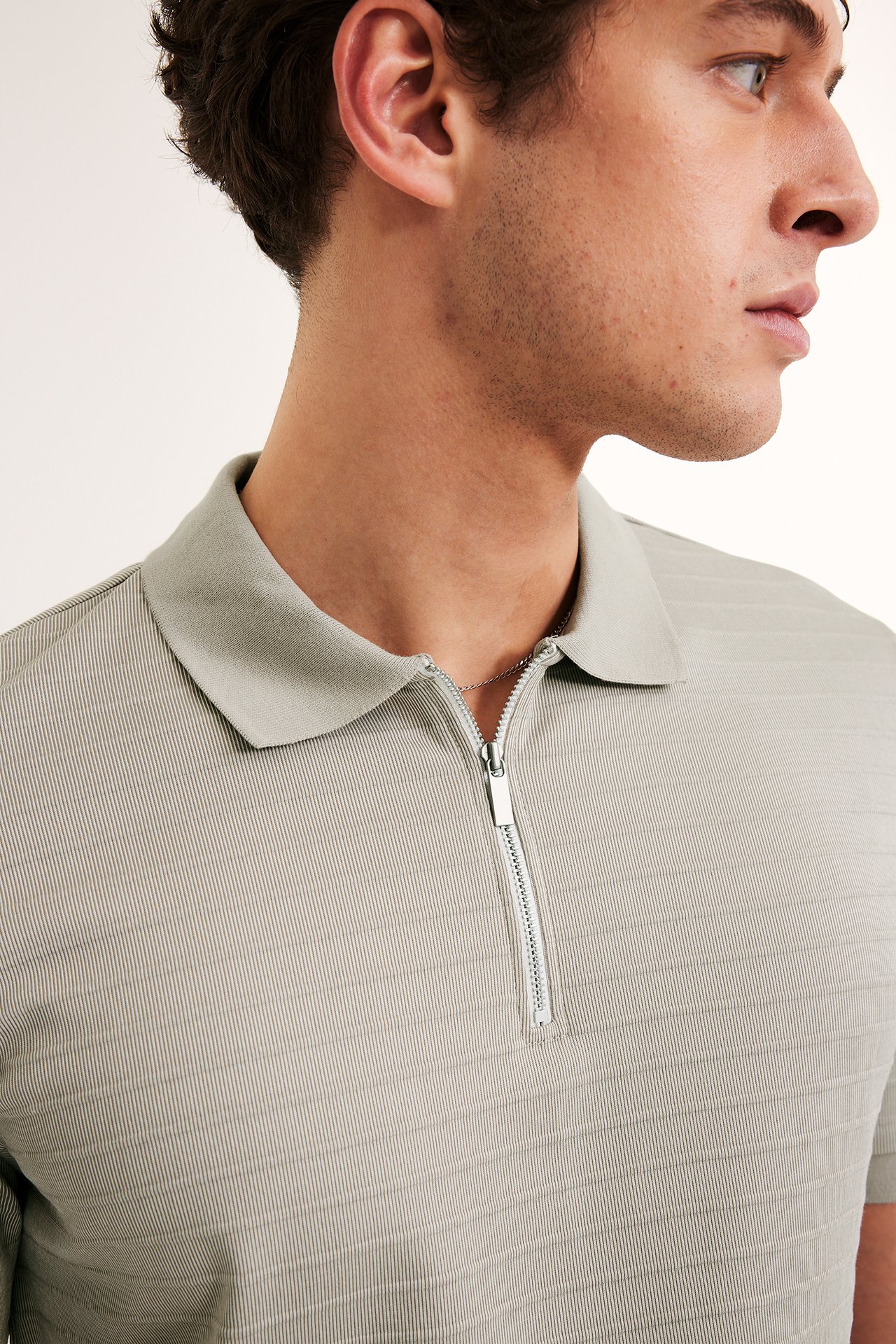Lähikuva miehestä sivulta, yllään beige pikeepaita kauluksella, hopeavetoketjulla ja teksturoituilla raidoilla.