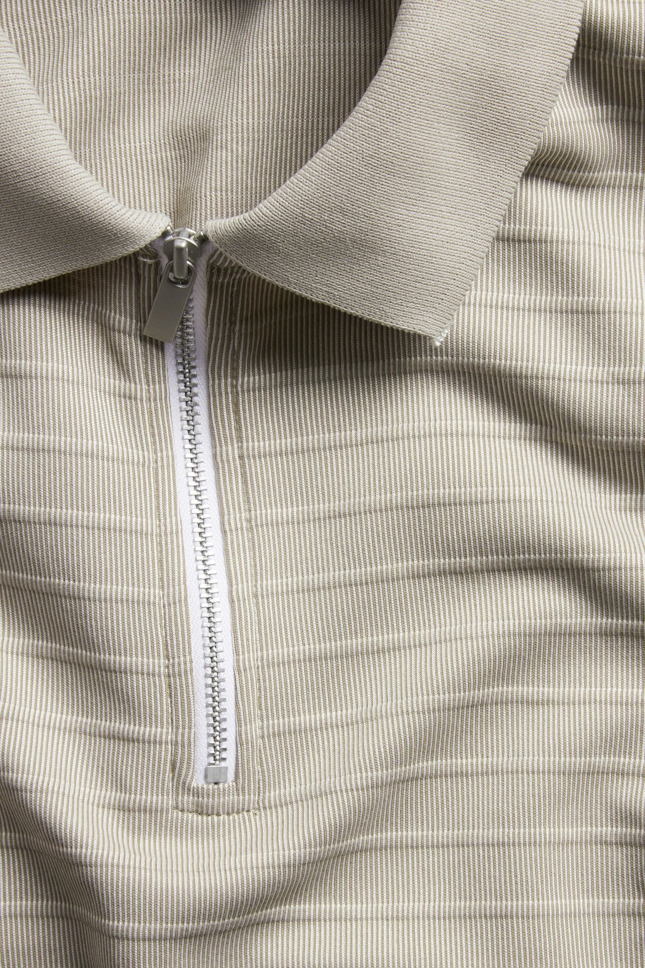 Nærbilde av en beige teksturert pikétrøye med pikékrage og hvit glidelås.