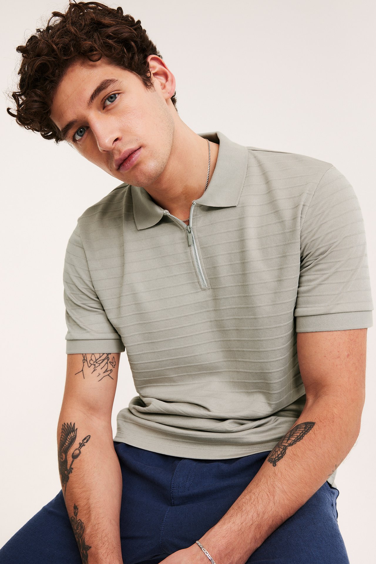 Mies yllään beige ribattu lyhythihainen pikeepaita, jossa pikeekaulus, vetoketju, ja siniset housut.