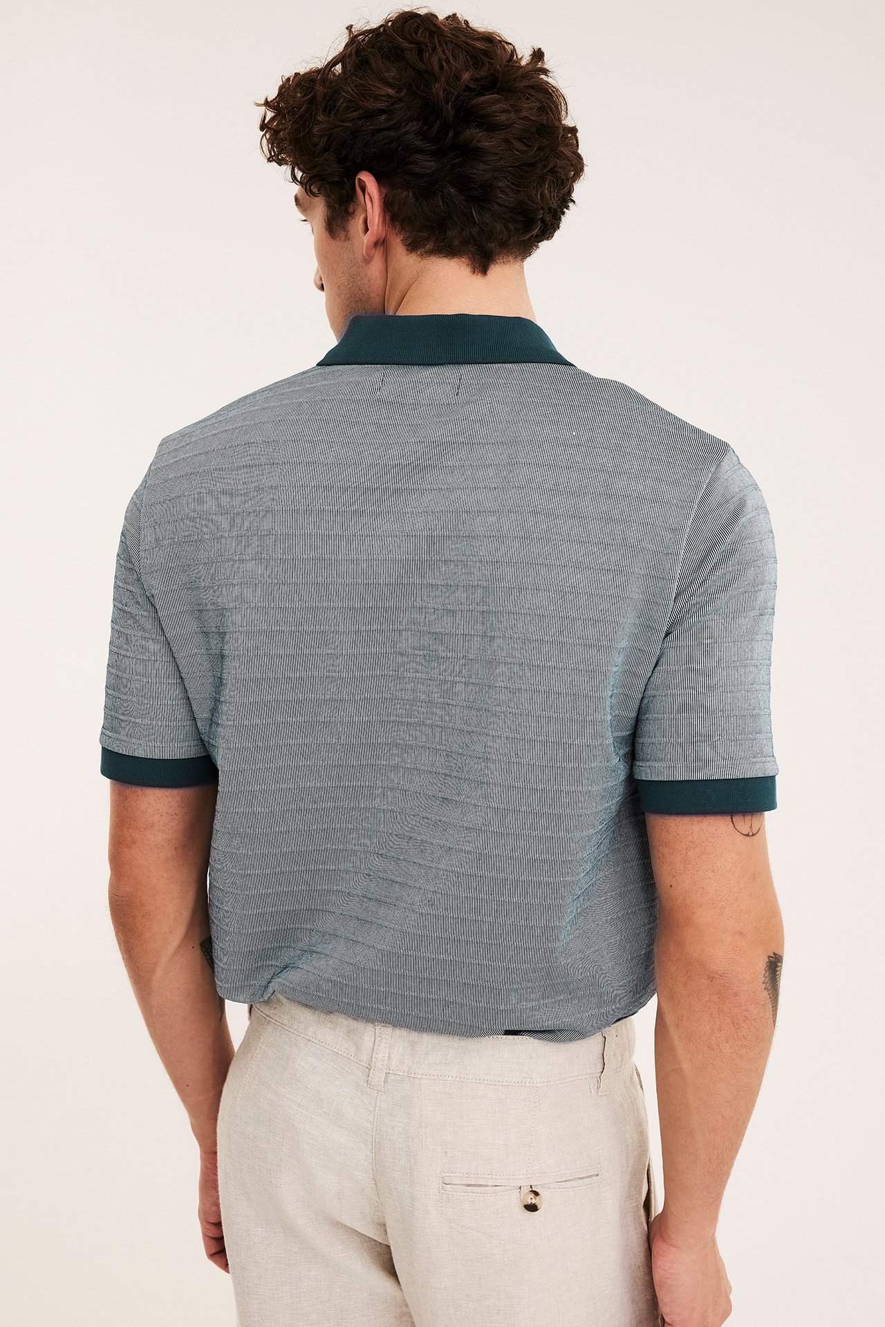 Mies takaa, yllään tummansinivihreä kuvioitu pikeepaita tummalla kauluksella ja hihansuilla, ja vaaleat beiget housut.