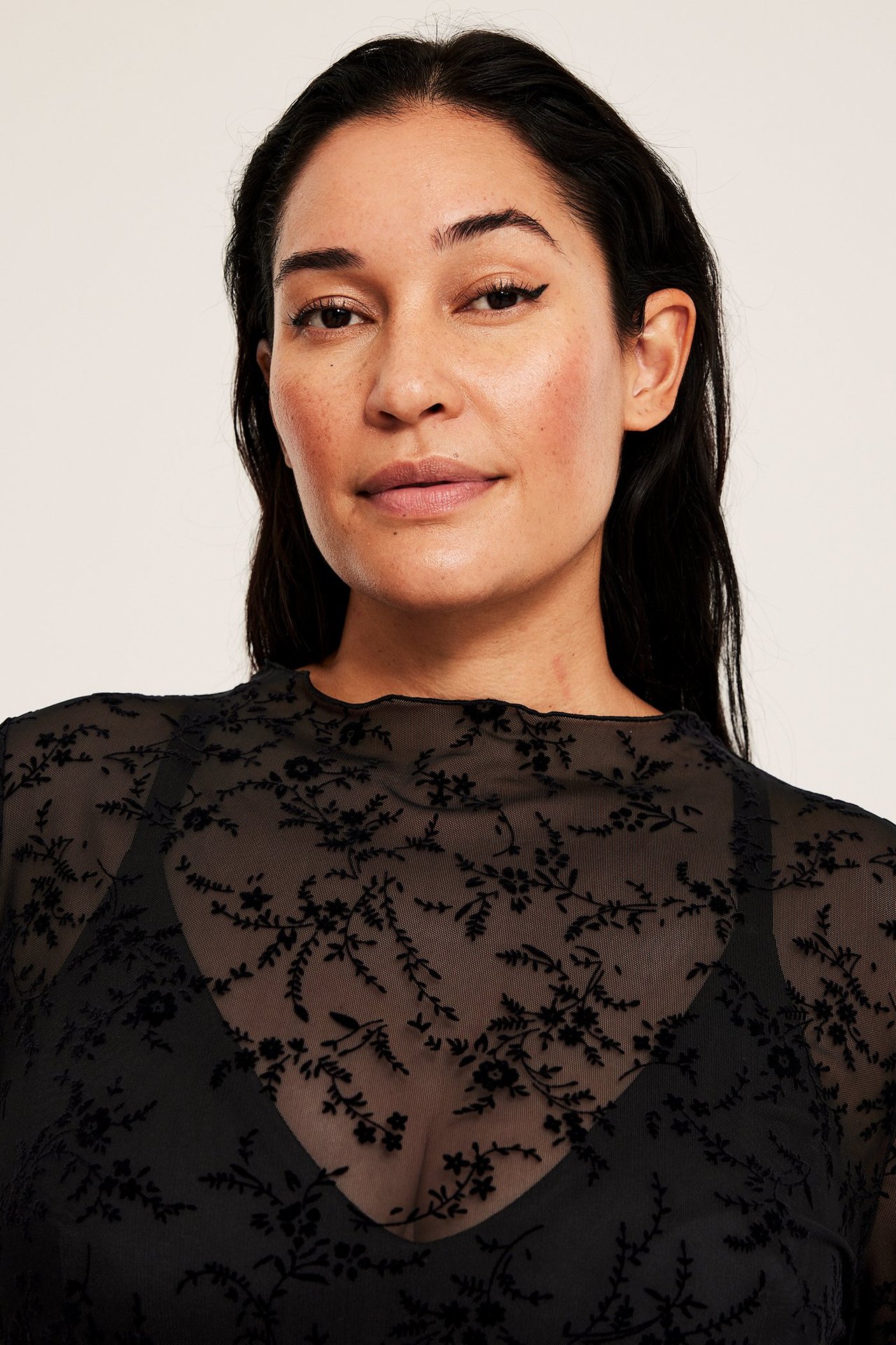 Närbild på kvinna i en svart, skir långärmad topp med flockat blommönster över svart BH.