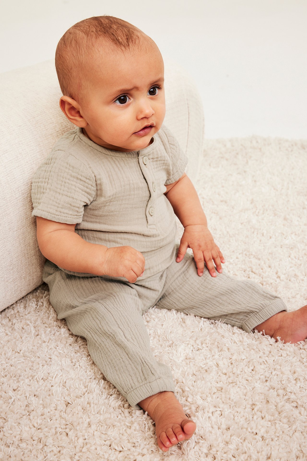 Frontvy av ett baby som bär ljusbeige muslinbyxor med resår i benslut och matchande kortärmad topp.