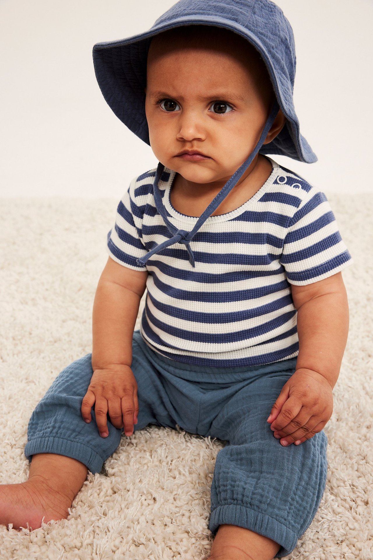Framifrån: baby i blå muslinbyxor, en vit- och blårandig tröja och en blå solhatt.