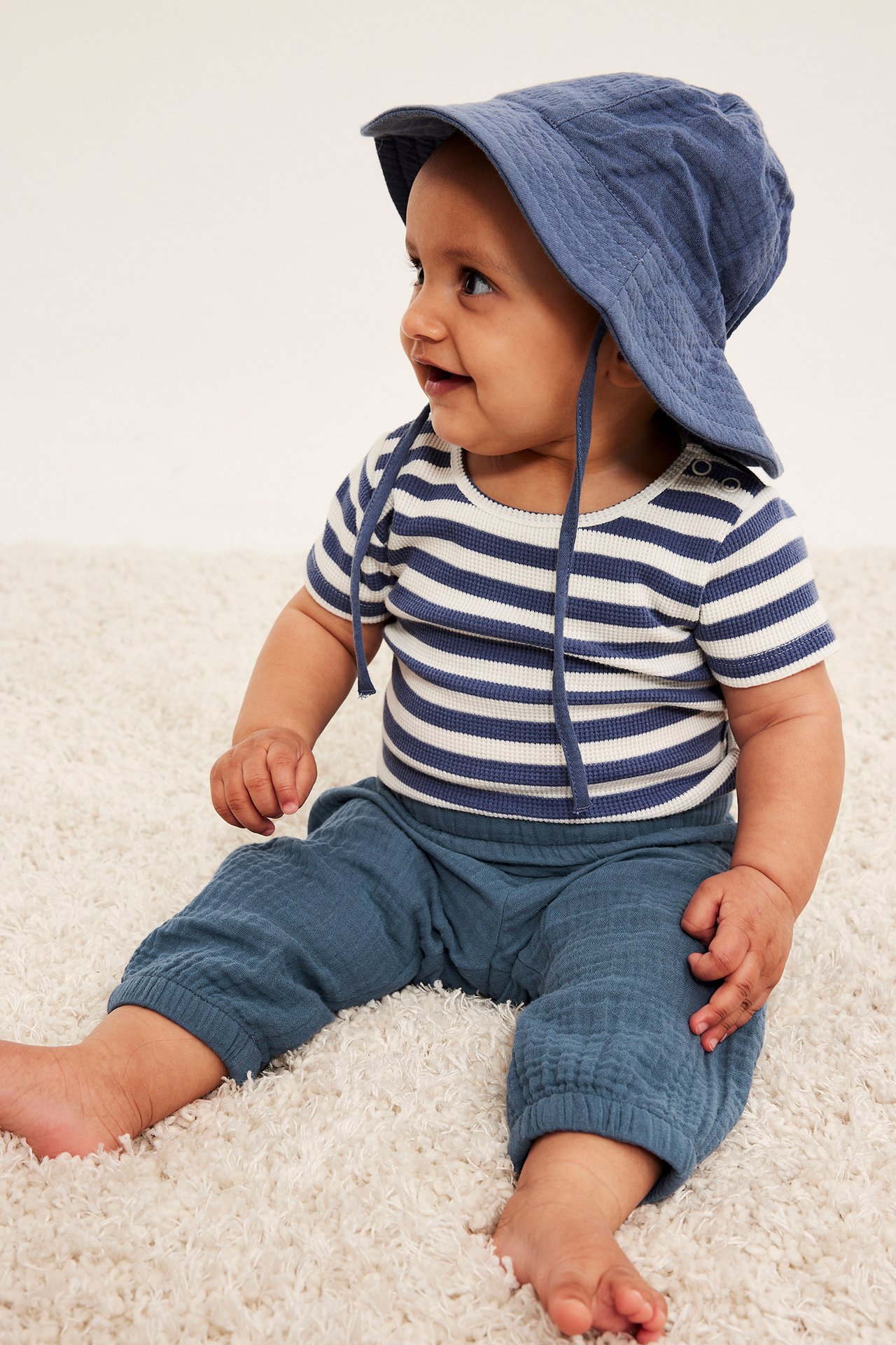 Baby bär blåa muslinbyxor med resår i benslut, en randig topp och en blå muslinhatt.