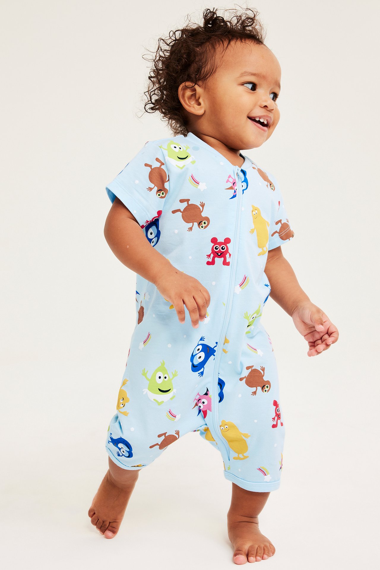 Edestä kuvattu vauva vaaleansinisessä kuvioidussa Babblarna-lyhythihaisessa haalarissa vetoketjulla.