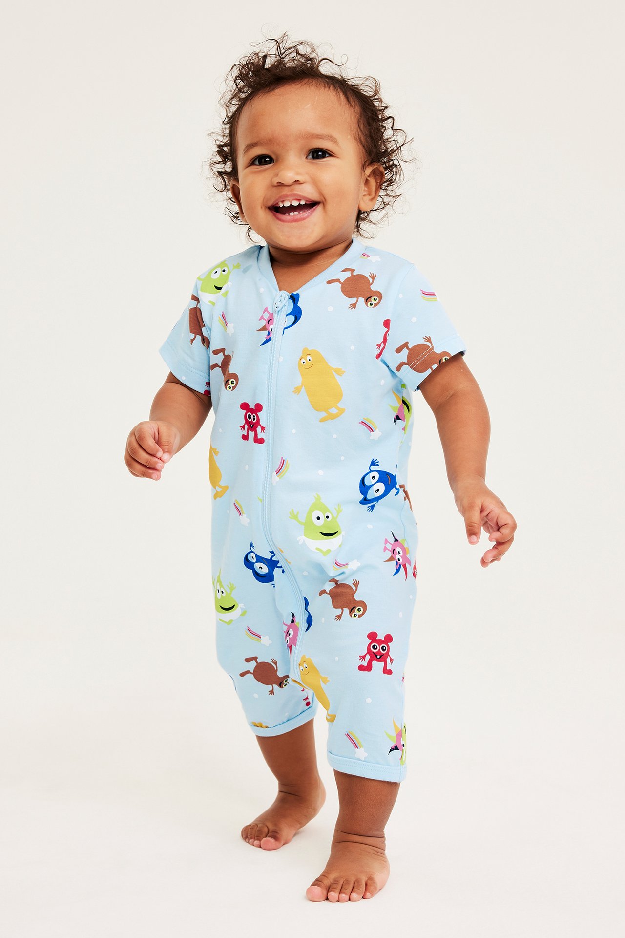 Edestä kuvattu iloinen vauva yllään vaaleansininen, kuvioitu, lyhythihainen haalari etuvetoketjulla.