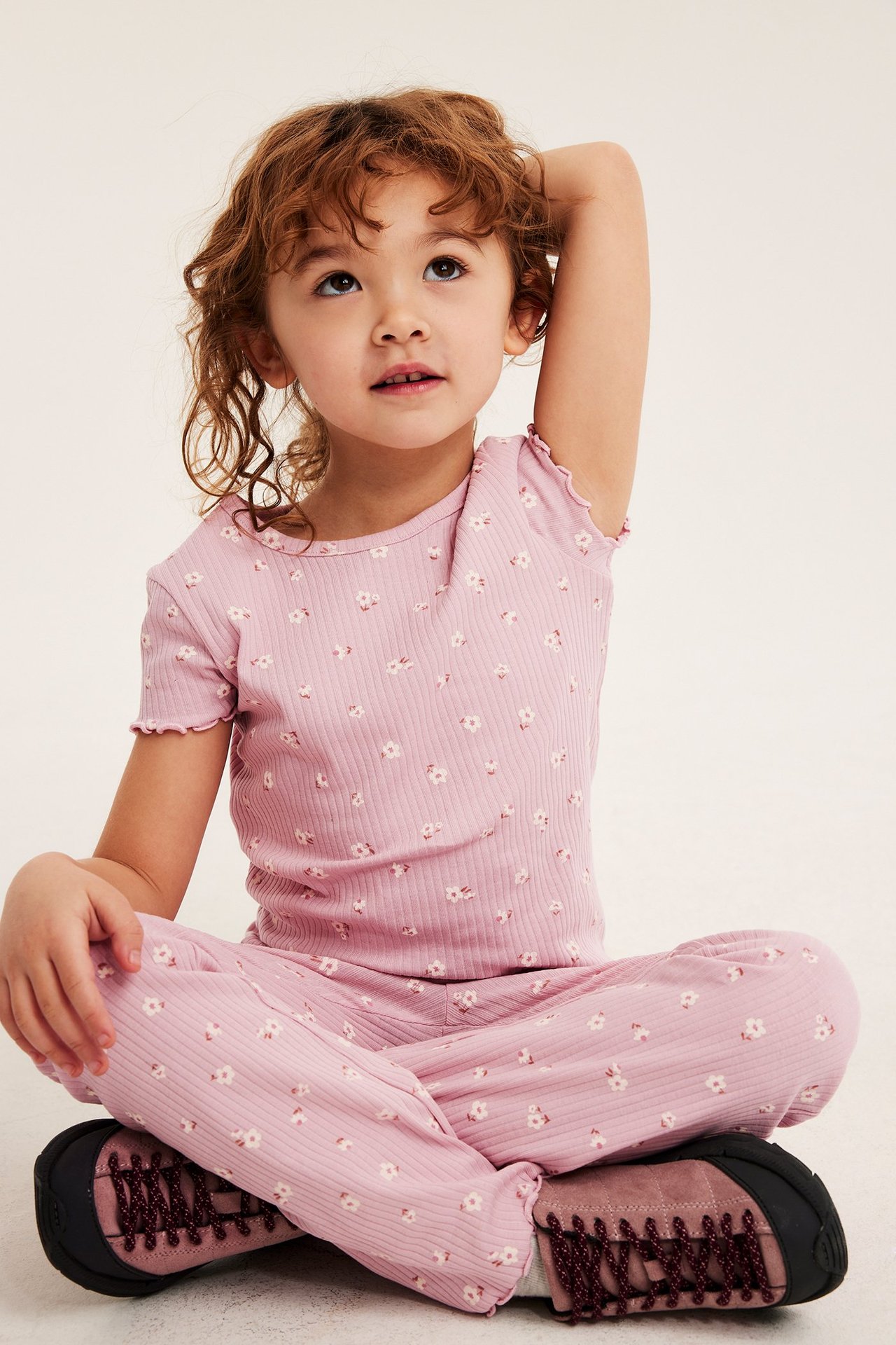 Edestä, lapsi vaaleanpunaisessa ribatussa lyhythihaisessa topissa ja yhteensopivissa kukkahousuissa, tummat kengät.