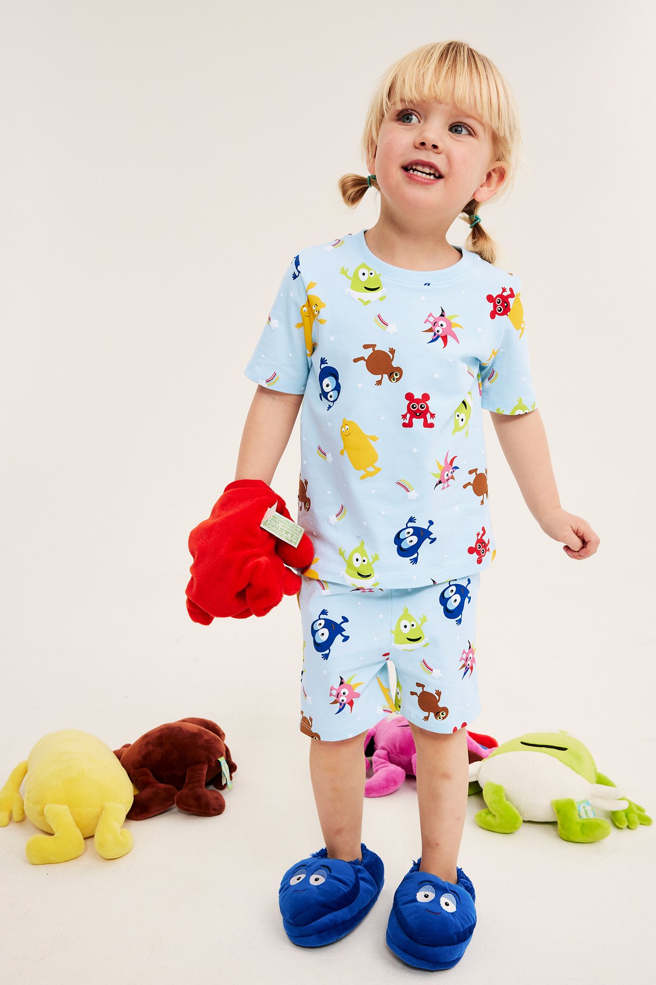 Framifrån: Ett barn i ljusblått Babblarna-mönstrat pyjamasset, håller en röd leksak och bär blå tofflor.