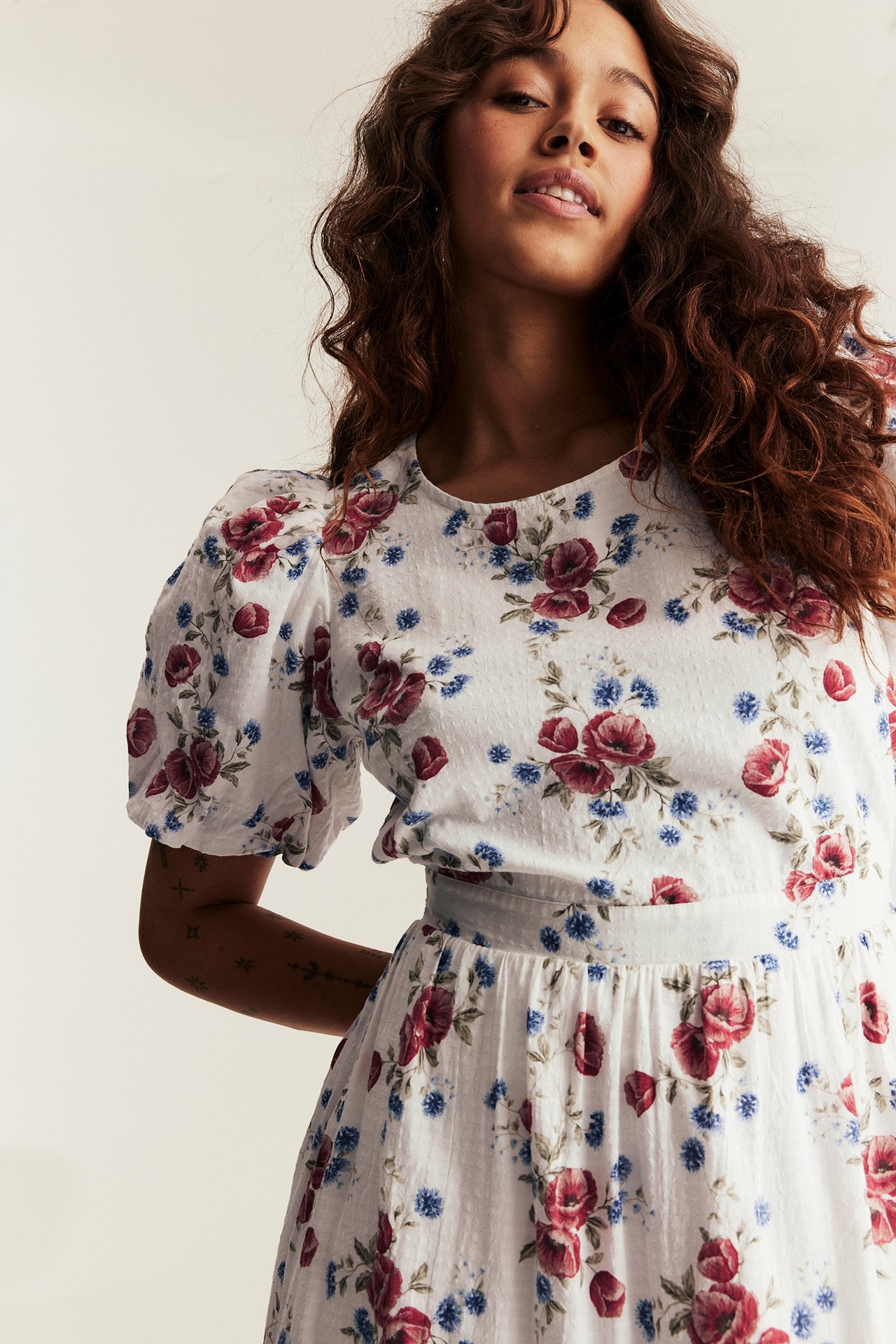 Frontvy: Kvinna i offwhite blommig klänning med röda och blå blommor, ballongärmar.