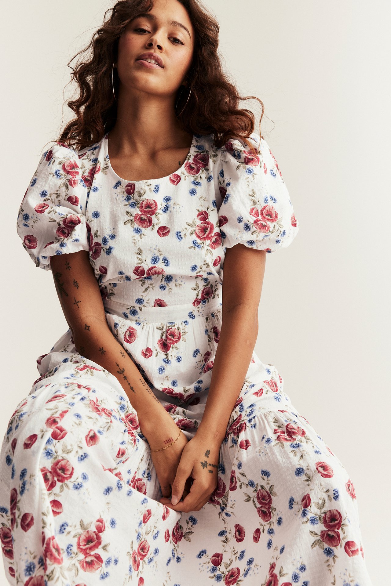 Edestä, istuva nainen yllään luonnonvalkoinen kudottu kukkamekko pussihihoilla ja pienillä rengaskorvakoruilla.