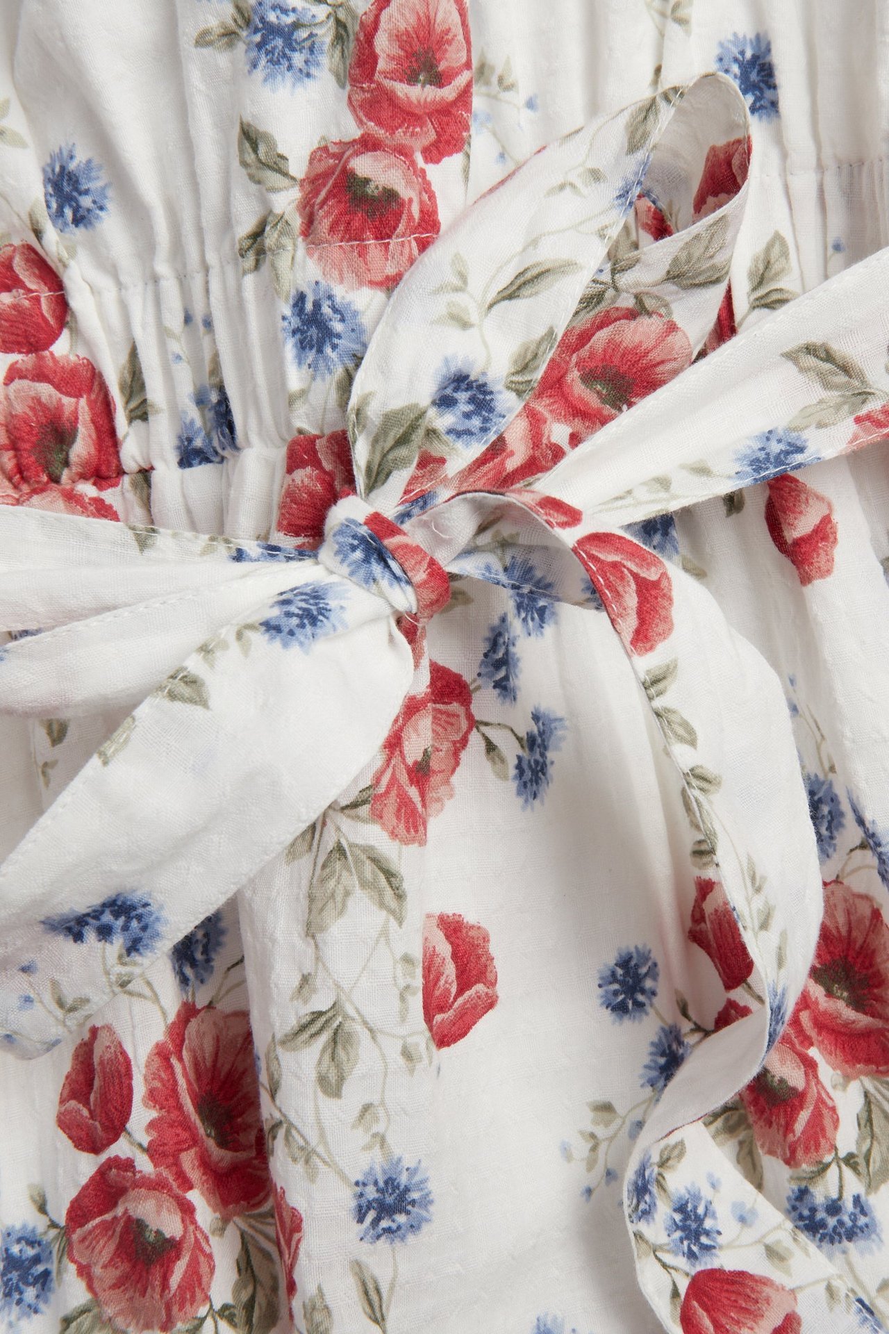 Lähikuva luonnonvalkoisesta kukkamekostä, jossa punaisia unikkoja ja sinisiä kukkia, rusetilla.