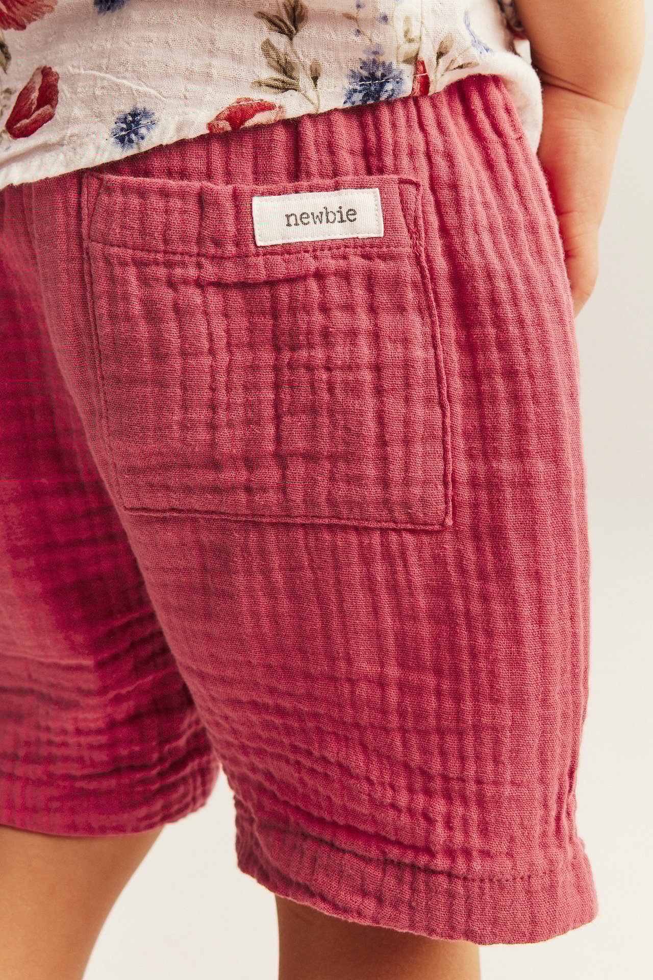 Närbild på barns texturerade röda vävda bomullsshorts med bakficka och Newbie-etikett.
