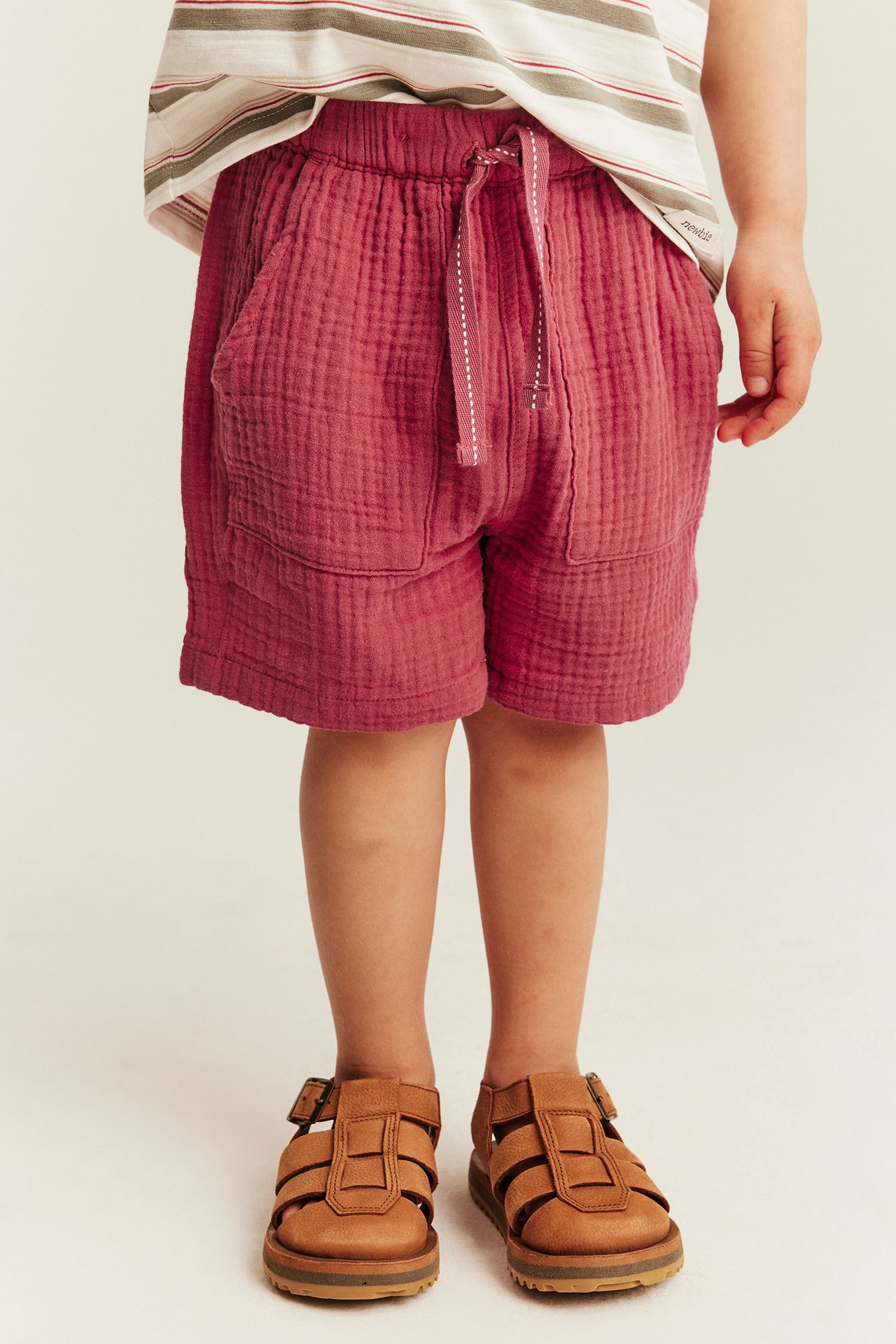 Widok z przodu, dziecko w czerwonych bawełnianych szortach ze sznurkiem, kieszeniami, pasiastym t-shirtem i sandałami.